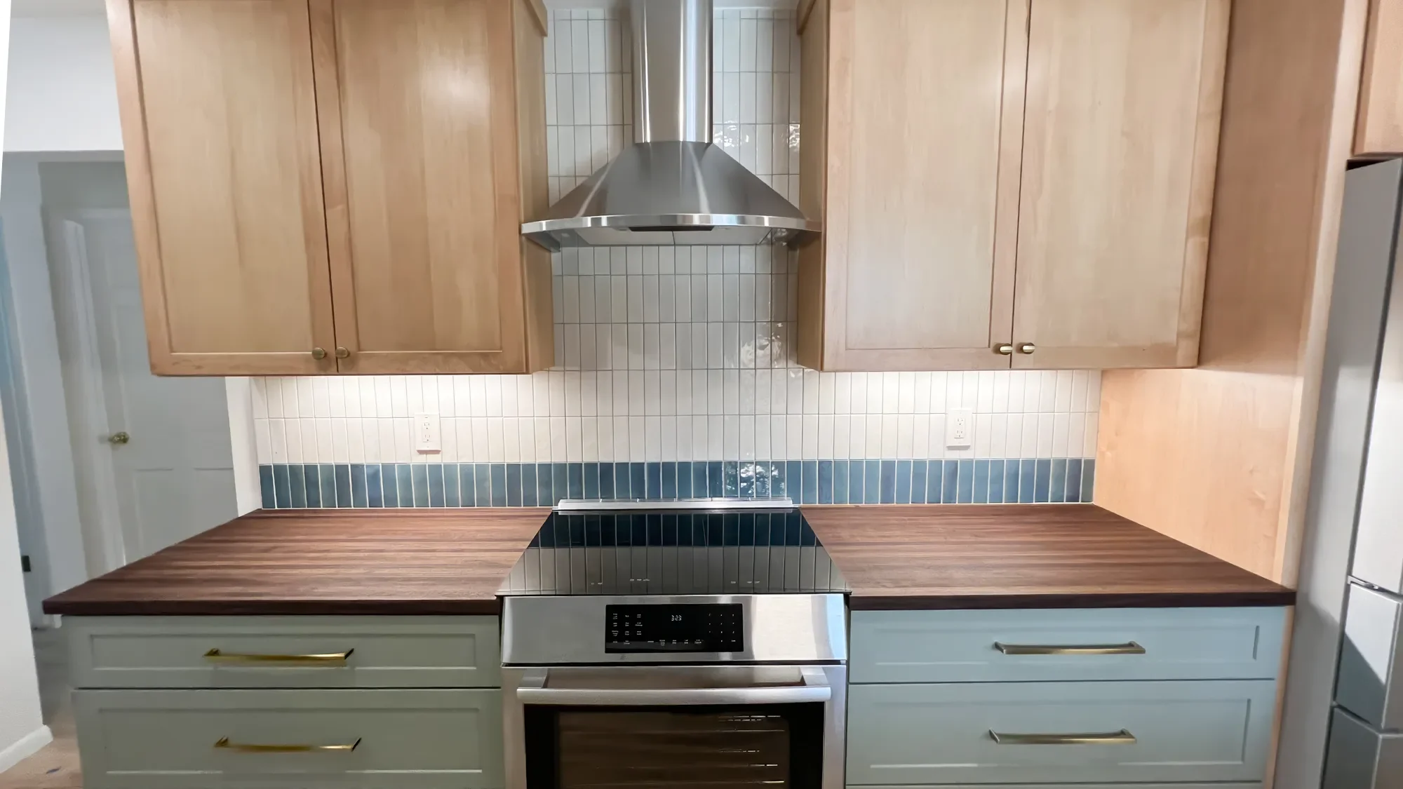Banner stove view of mixed-material transitional craftsman kitchen remodel in Longmont, CO featuring stainless steel stove and range hood, natural maple and sage green cabinets, dark walnut countertops, and white and teal full height tile backsplash