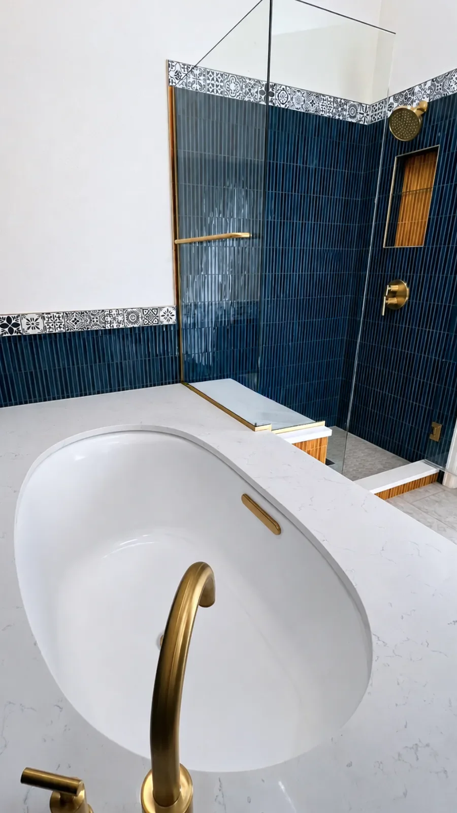 View of oval Koehler soaking tub and frameless glass walk-in shower with quartz top bench seating, gold fixtures, blue shower tile and wainscoting, and a gold tiled shelf shower niche in a Gunbarrel, CO luxury bathroom remodel