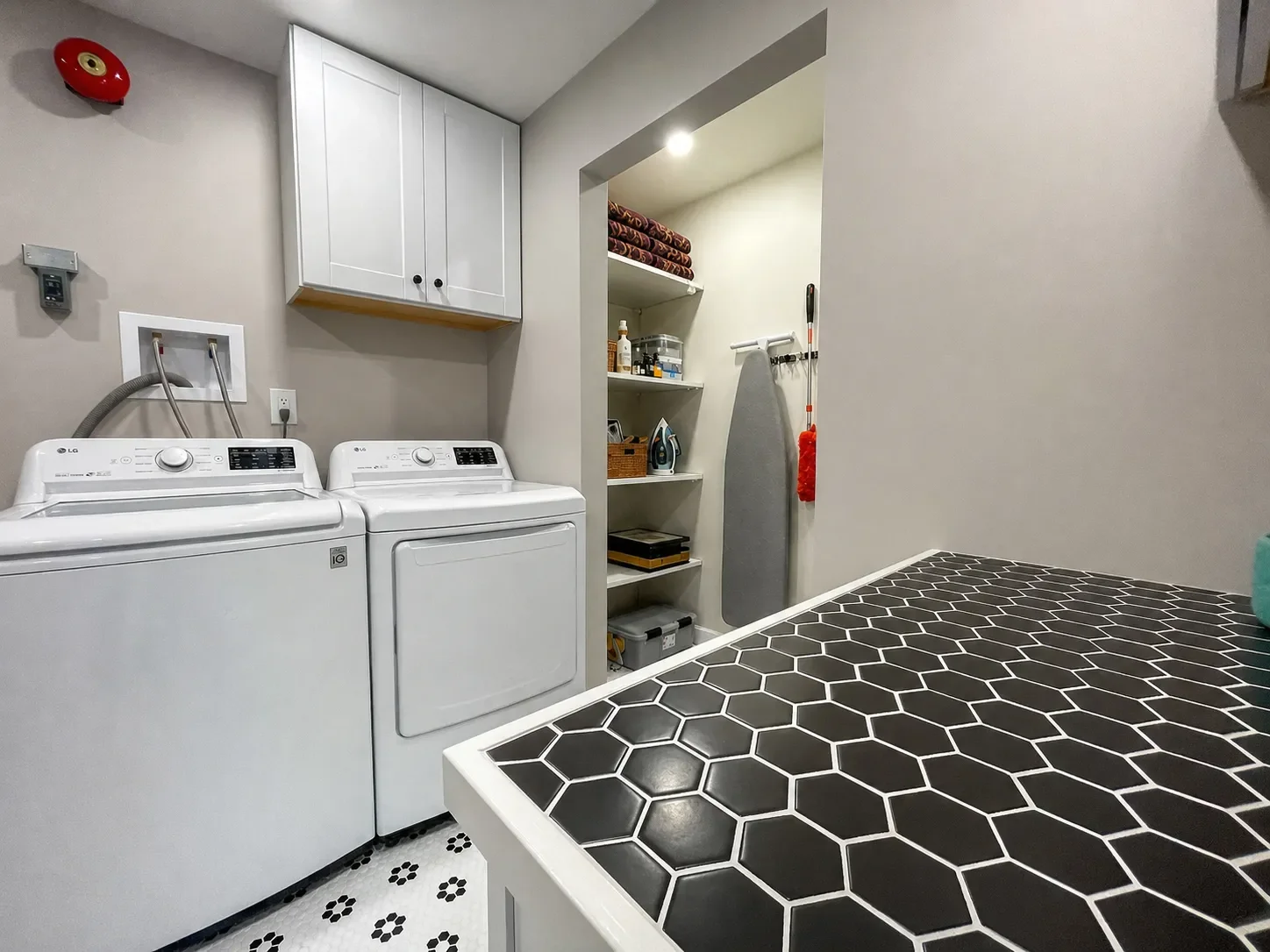 Laundry room with a washing machine and dryer, overhead cabinets, black hexagon tile counter, and a storage area with shelves and an ironing board.