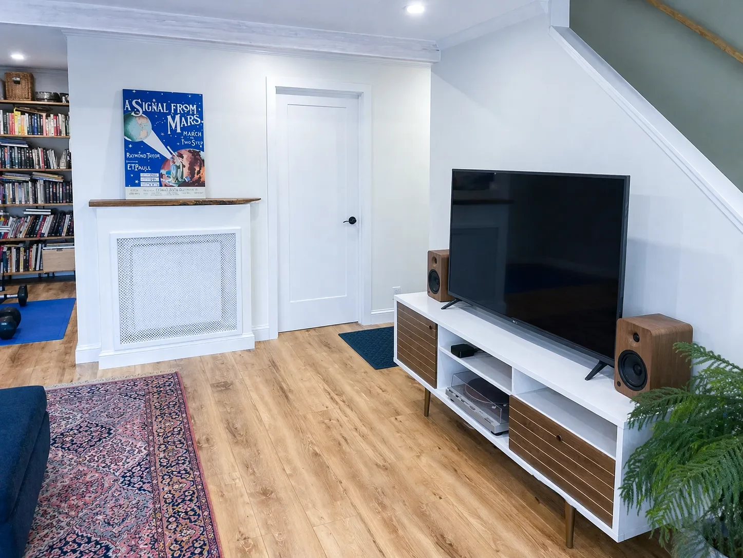 Living room with hardwood floors, a large TV on a white stand, bookshelf filled with books, and a movie poster. A patterned rug and a potted fern are present, next to a staircase with a wooden railing.