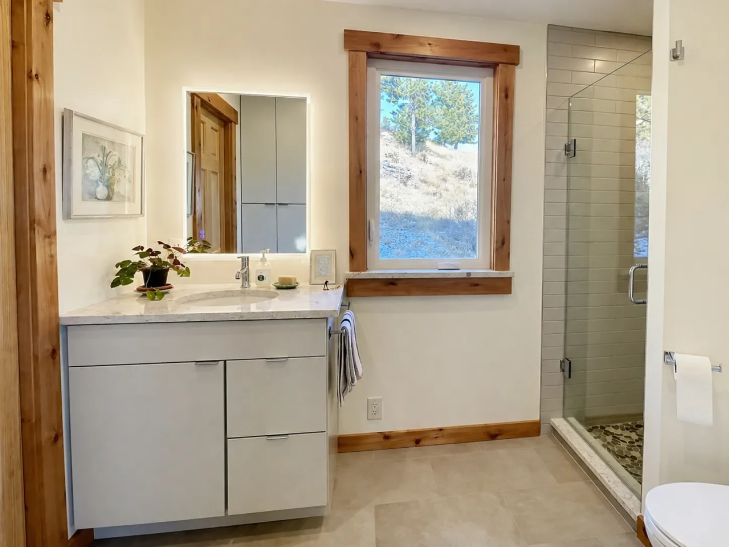 Rustic mountain bathroom remodel in Pine Brook Hills, Boulder, CO featuring euro-style gray vanity, quartz countertop, heated tiled floor, LED mirror, and wood trimmed window, door, and baseboards