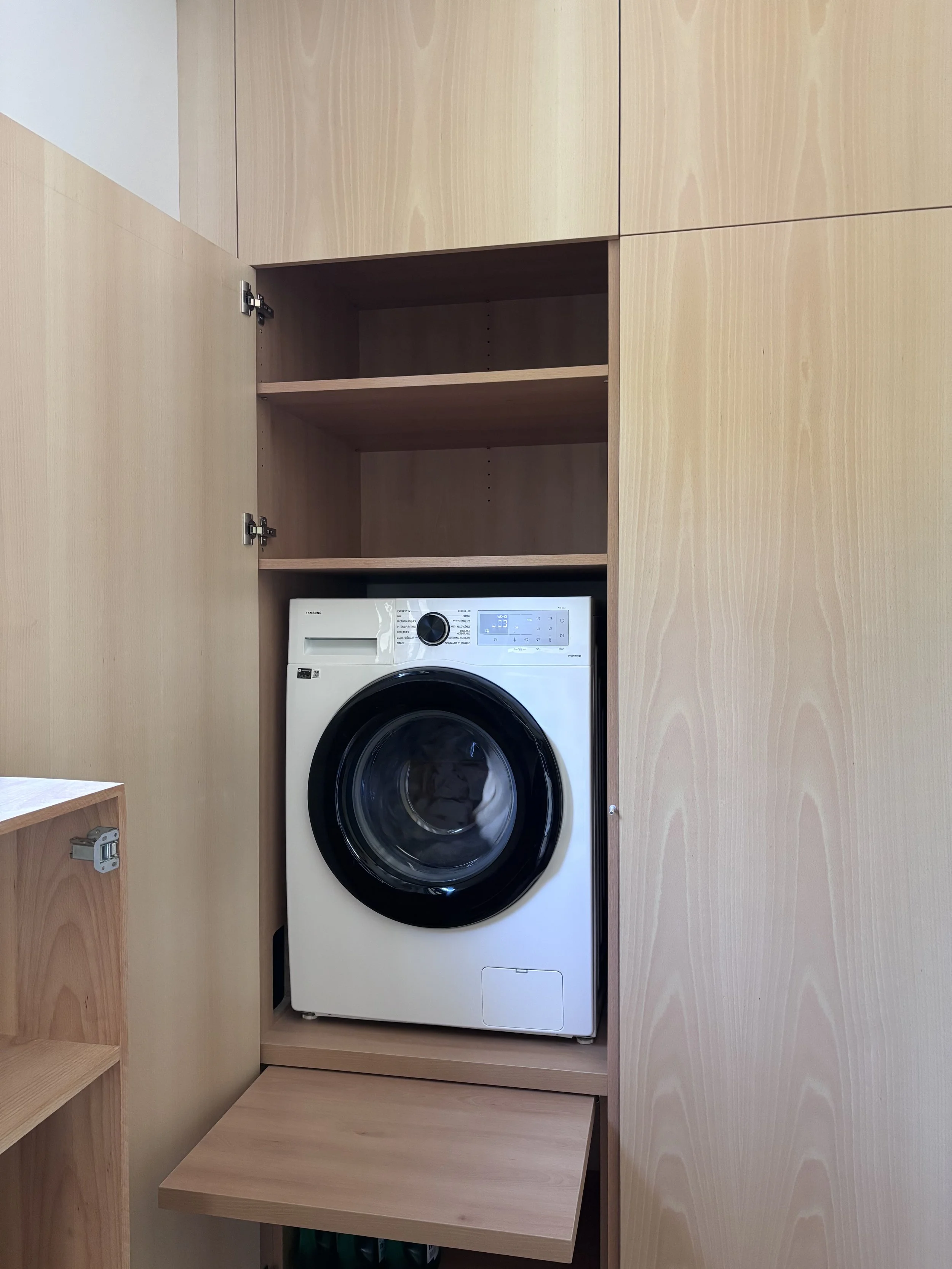 Lave-linge blanc intégré dans une armoire en bois clair, avec des étagères au-dessus.