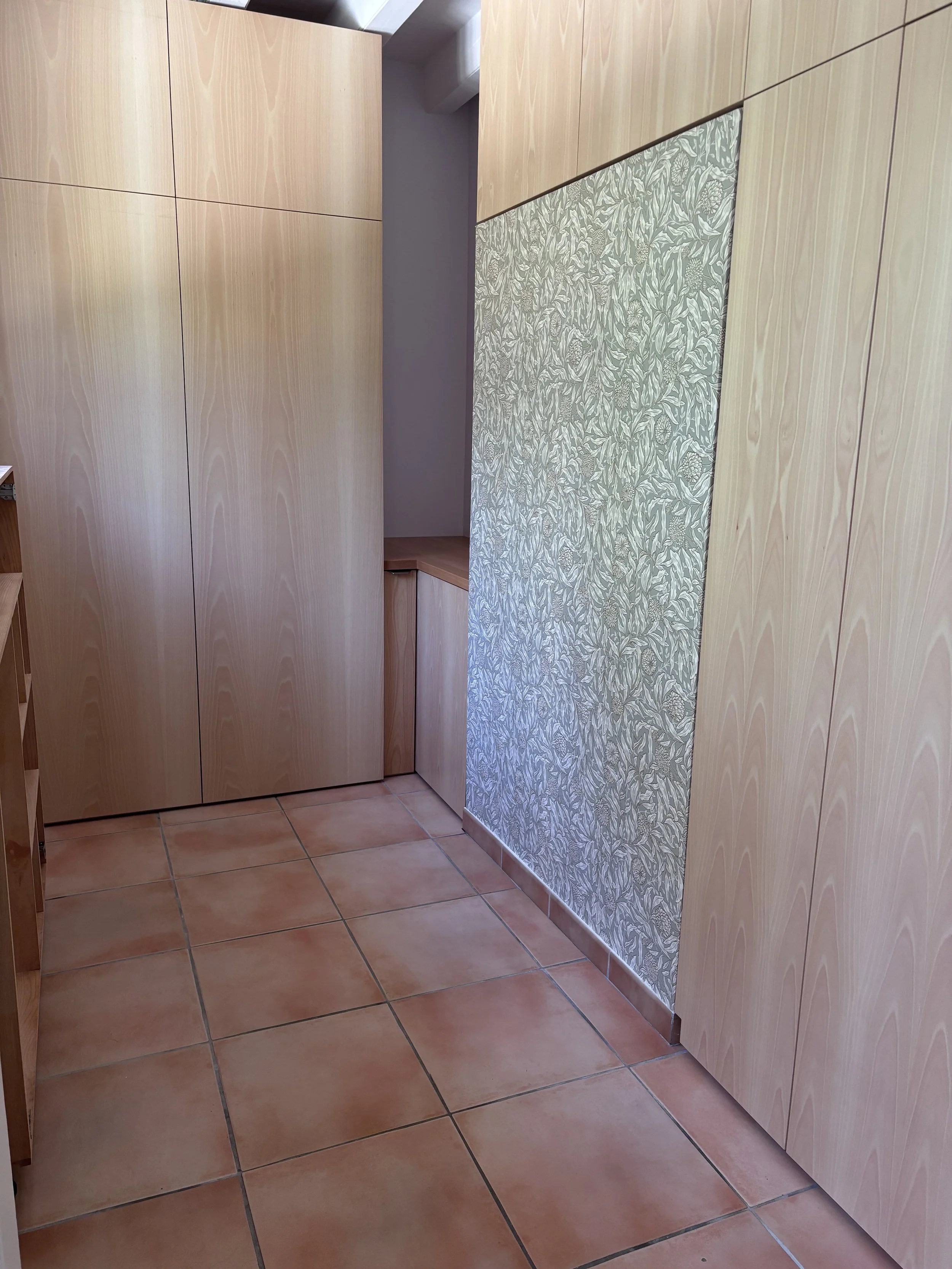 Petite pièce avec murs en bois clair, un mur partiellement recouvert de papier peint à motif végétal, sol en carreaux de terre cuite.