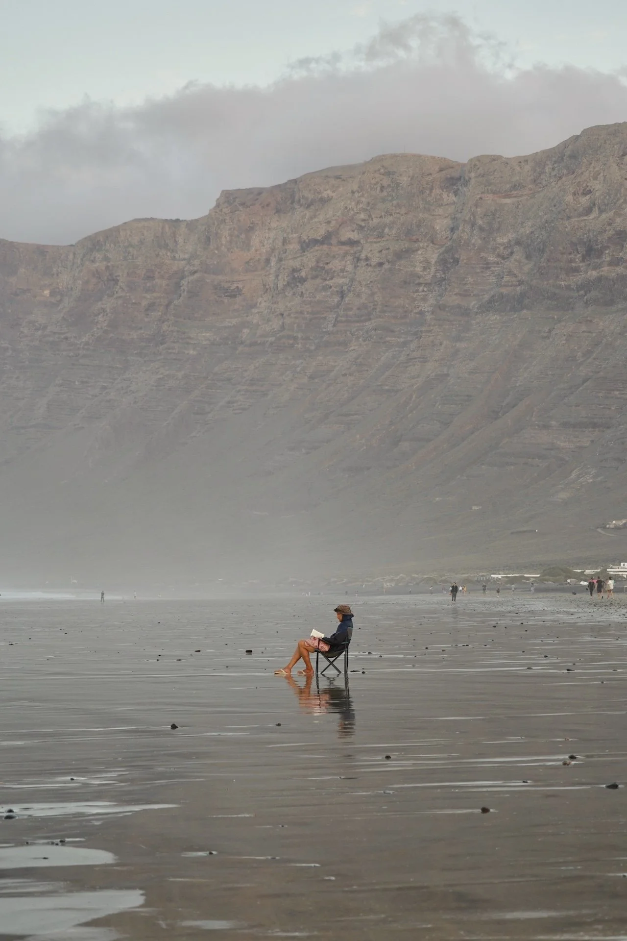 Person sitting in a chair reading a book on Famara Beach, Lanzarote, near the shoreline with mountains in the background.