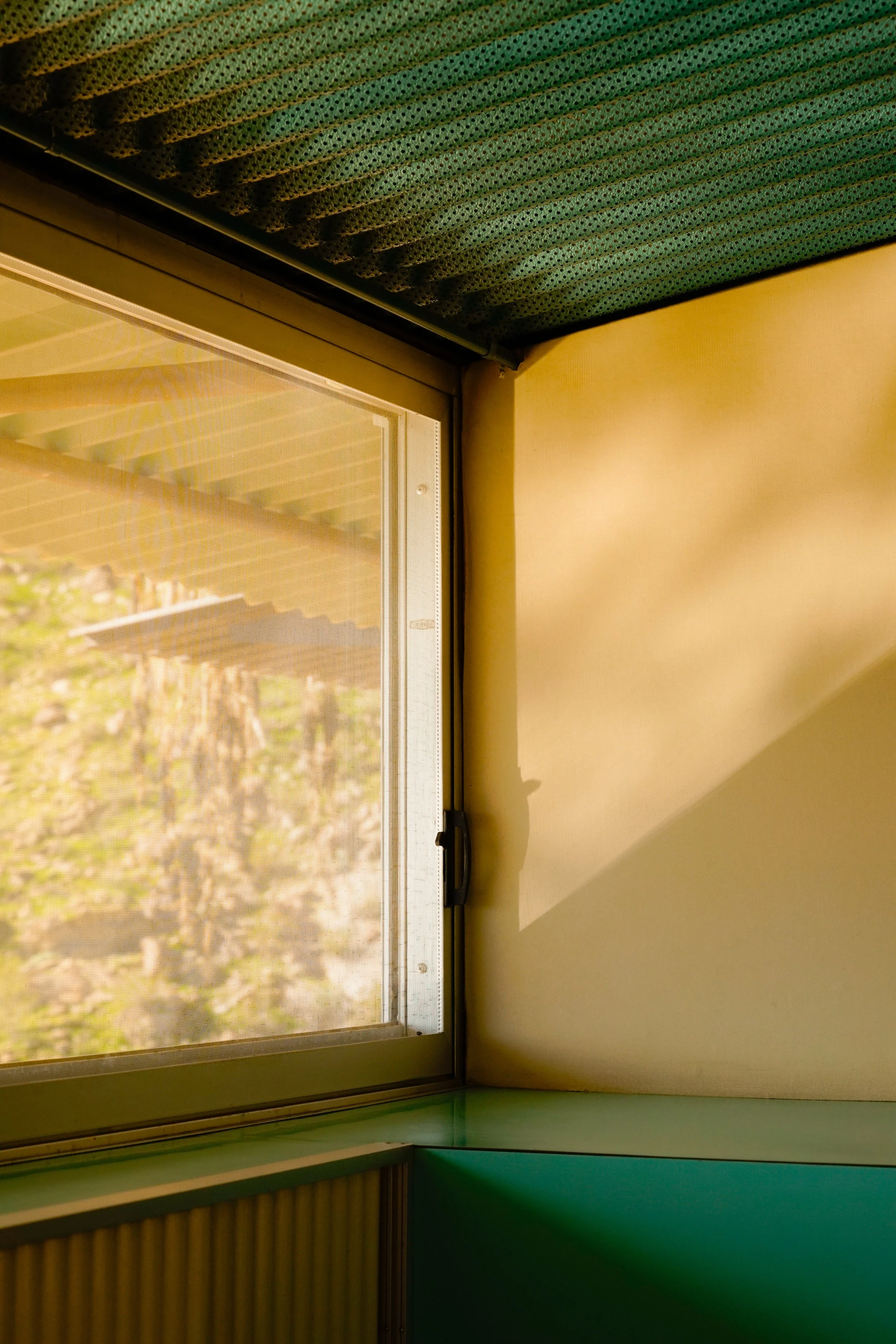 Interior view of a room with a large window or glass door, a green and yellow paper or fabric covering at the top, and a yellow wall on the right side. The window shows an outdoor scene with trees and sky.