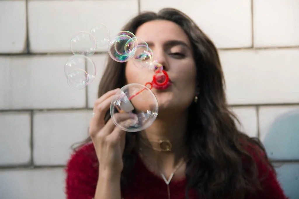 A woman with dark hair in a red sweater blowing bubbles with a bubble wand against a brick wall background.