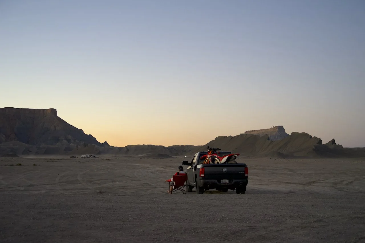 A person sitting in a red camping chair next to a black pickup truck in a desert with mountains in the background during sunset.