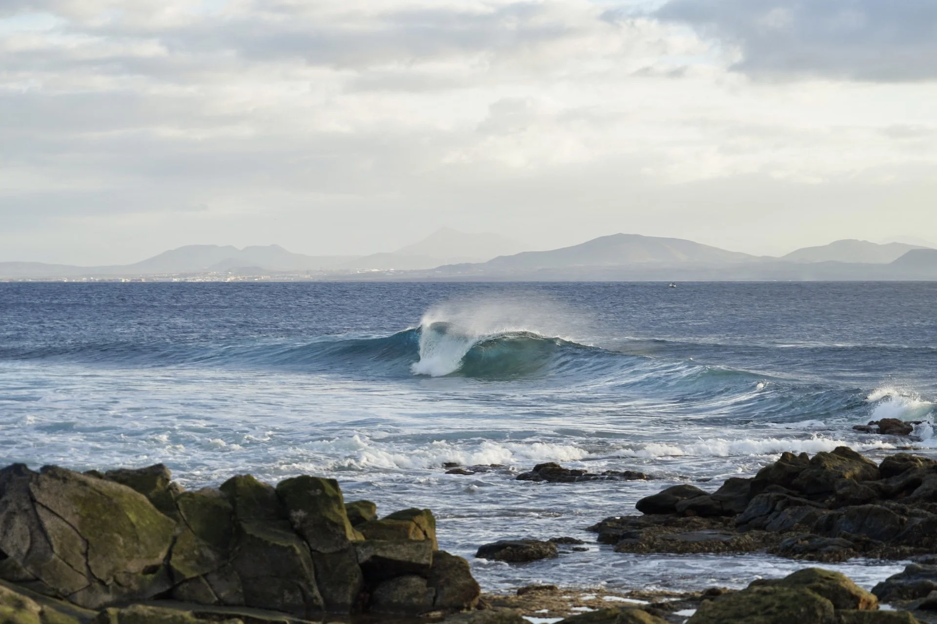 Ocean waves crashing against rocks on a shoreline with distant mountains and cloudy sky in the background.