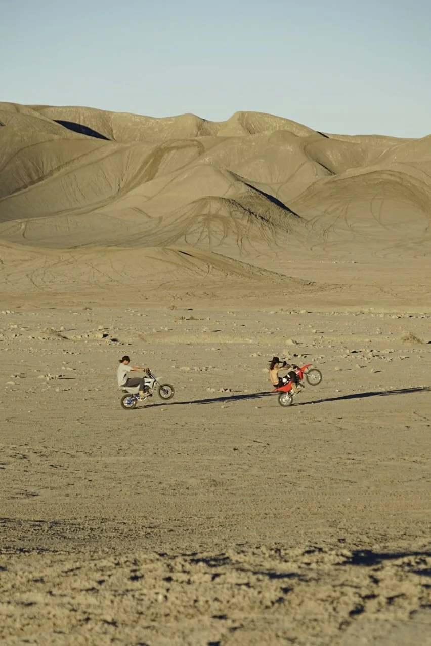 Two people riding dirt bikes near Factory Butte in Hanksville, Utah, surrounded by a desert landscape with sandy dunes.