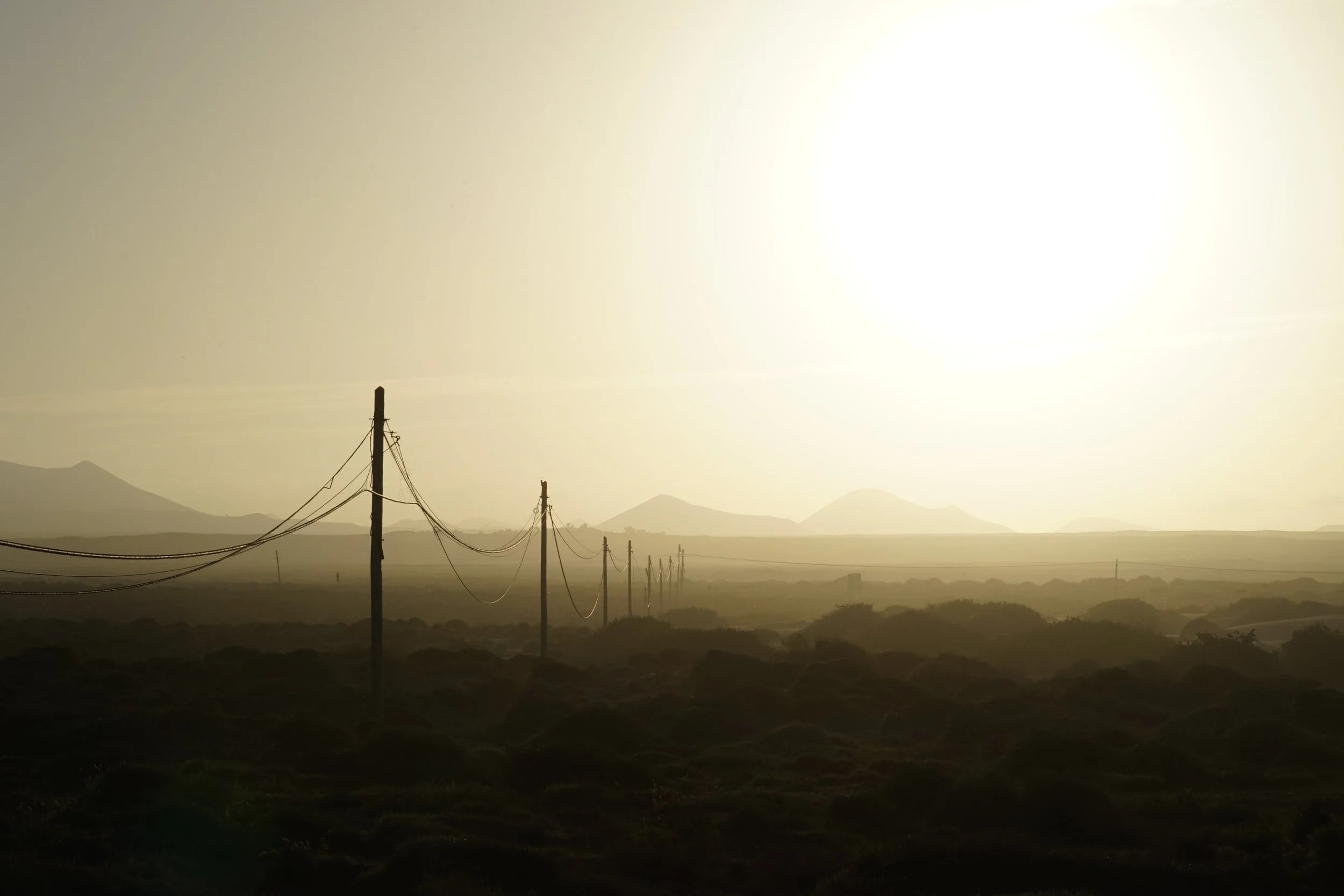A landscape scene of a dry area near Famara, Lanzarote, with multiple power poles and wires, mountains in the distance, and a hazy, bright sky.
