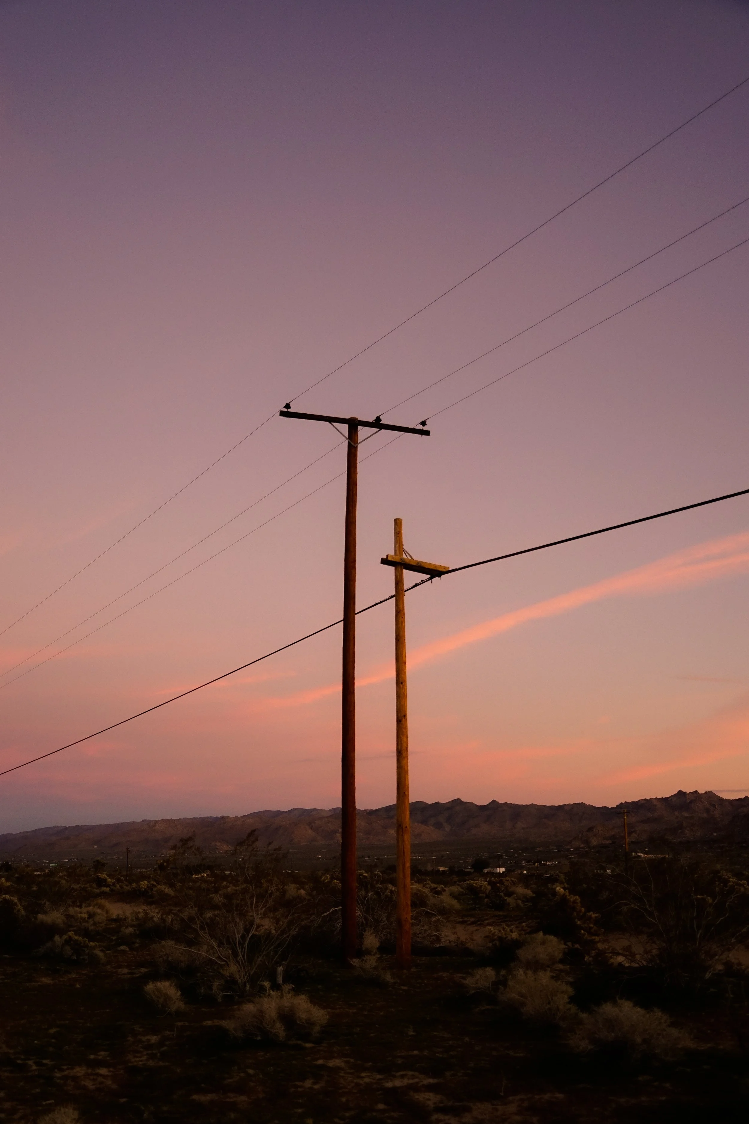 Two utility poles with power lines against a colorful sunset sky over a desert landscape with hills in the distance.