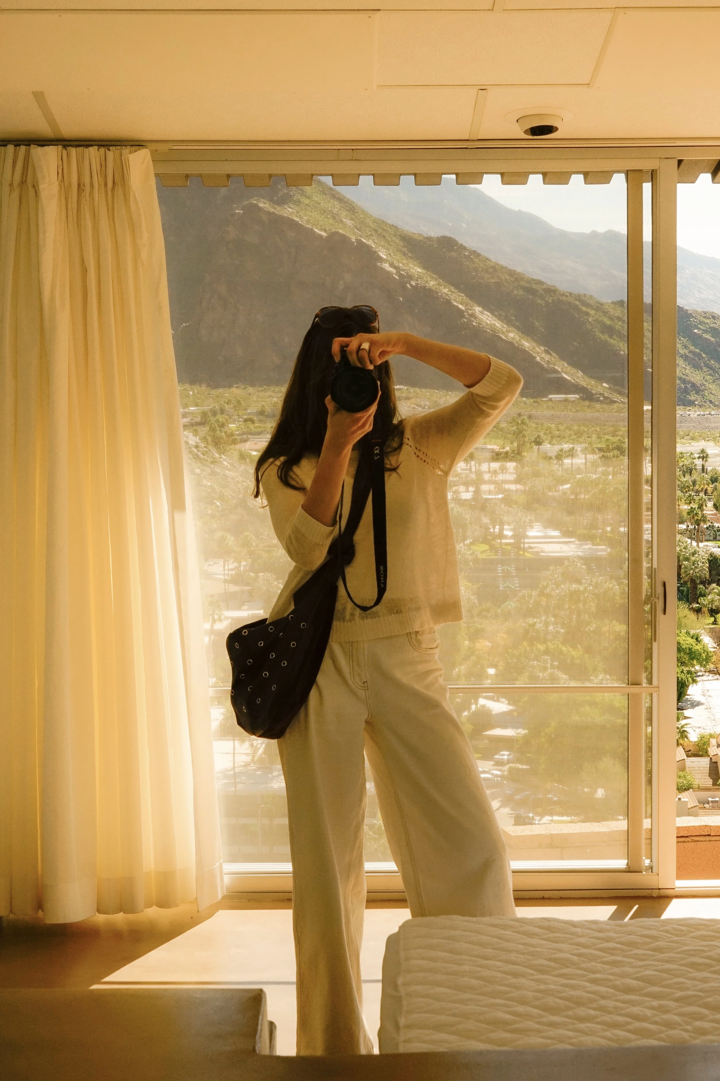A woman taking a mirror selfie inside a room with a large window, beige curtains, and a mountainous landscape outside.