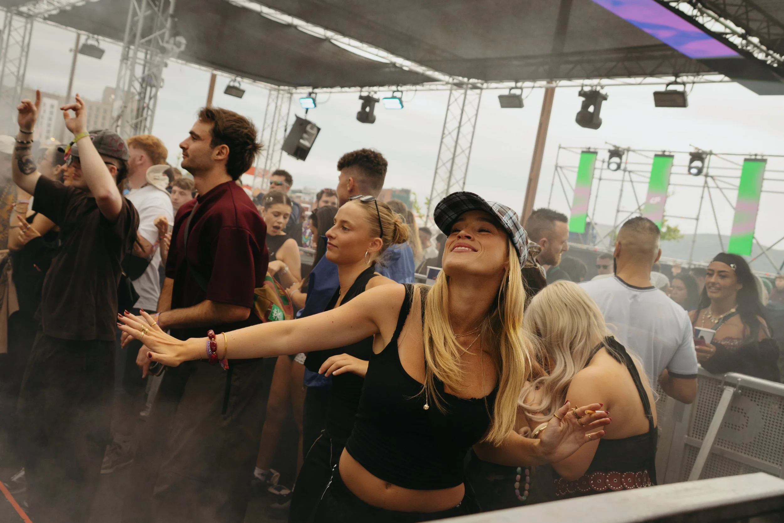 A crowd of people at the RA Stage at AVA music festival in Belfast, dancing and enjoying the event under a large stage with lights and screens, with some people smiling and others focused on the music.