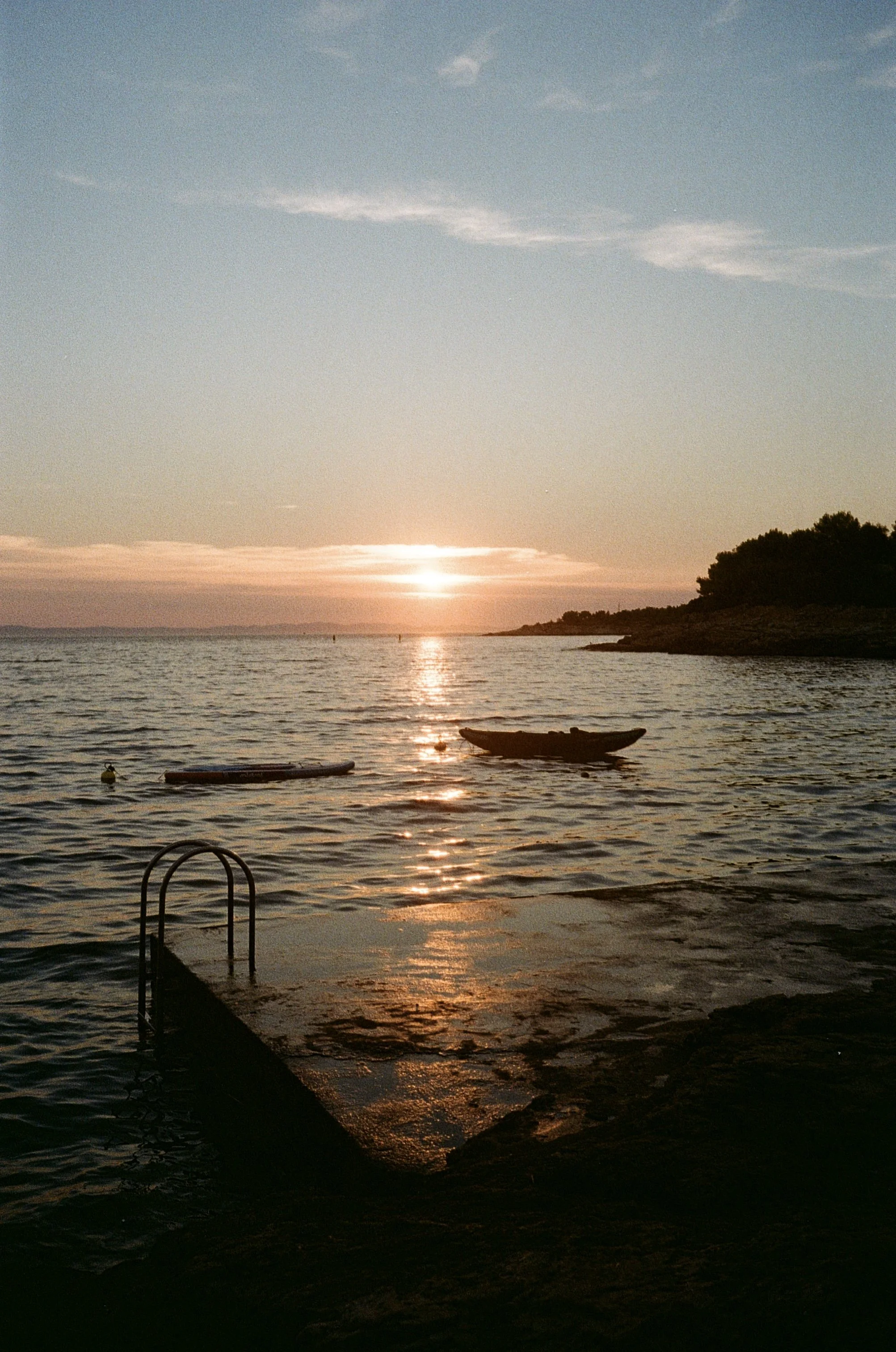 A sunset at Murter Island, Croatia, over a calm body of water with boats floating, a dock with a ladder in the foreground, and trees on the right side of the shoreline.