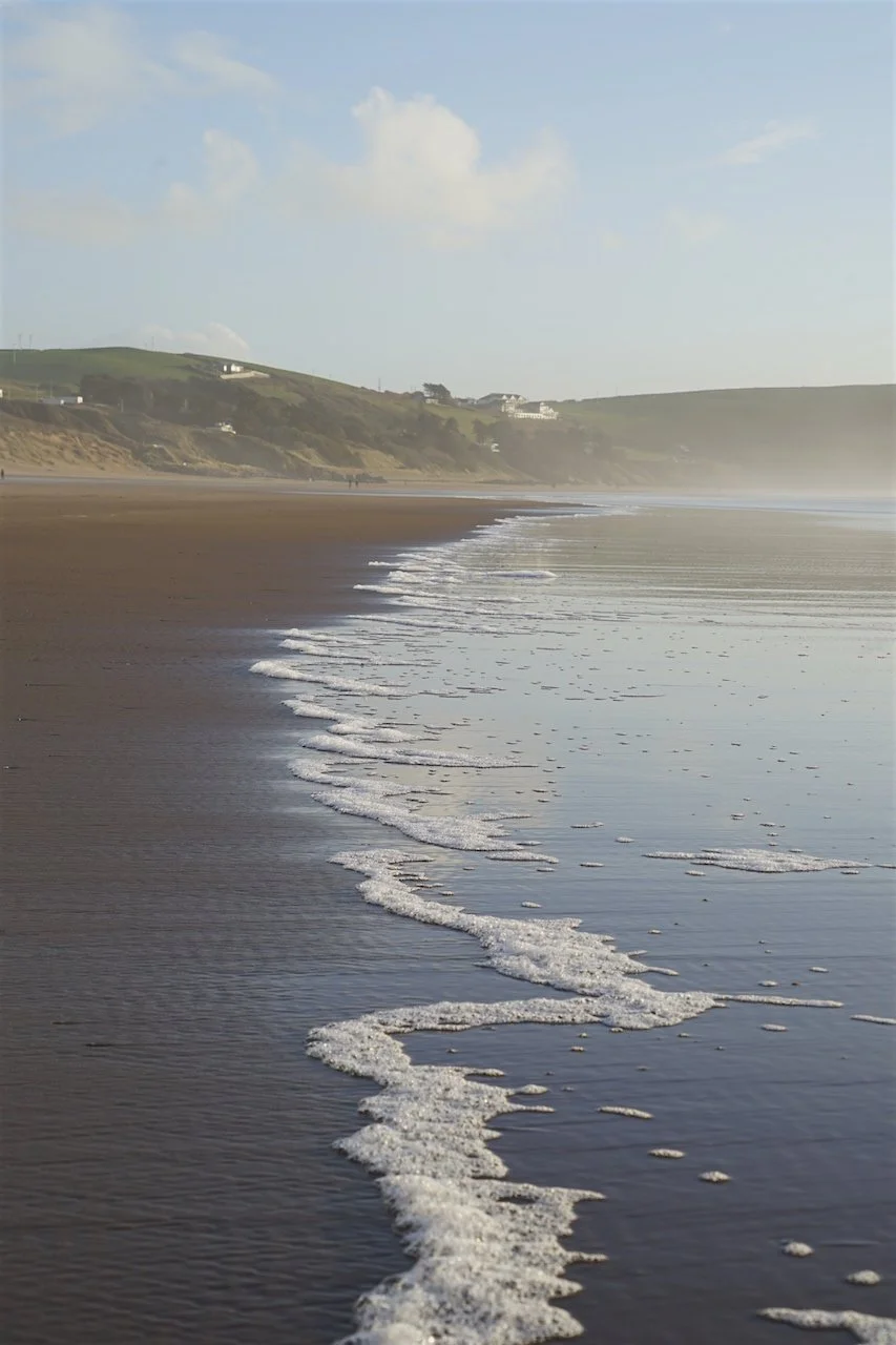 Woolacombe Beach in Devon, United Kingdom, showing reflections along the shoreline and natural foam patterns in the water.