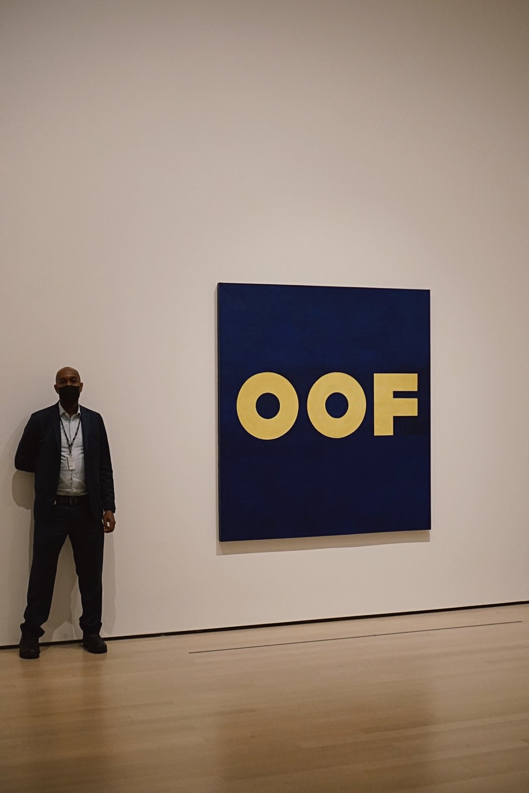 A security guard wearing a suit and face mask standing beside an Edward Ruscha painting titled “OOF” at MoMA in New York City.