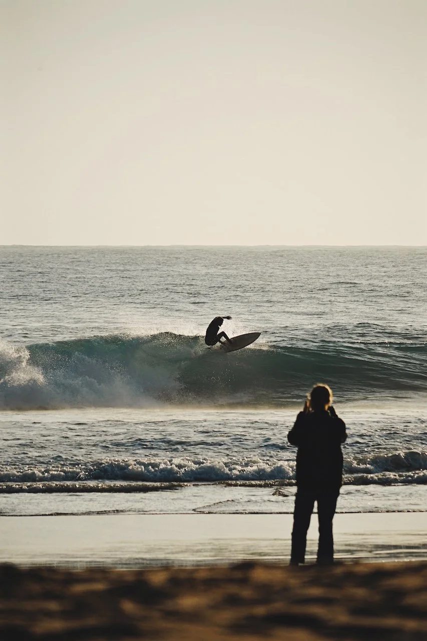 A surfer riding waves at sunset in Sagres, Portugal, with a woman standing on the beach watching the ocean and taking pictures.