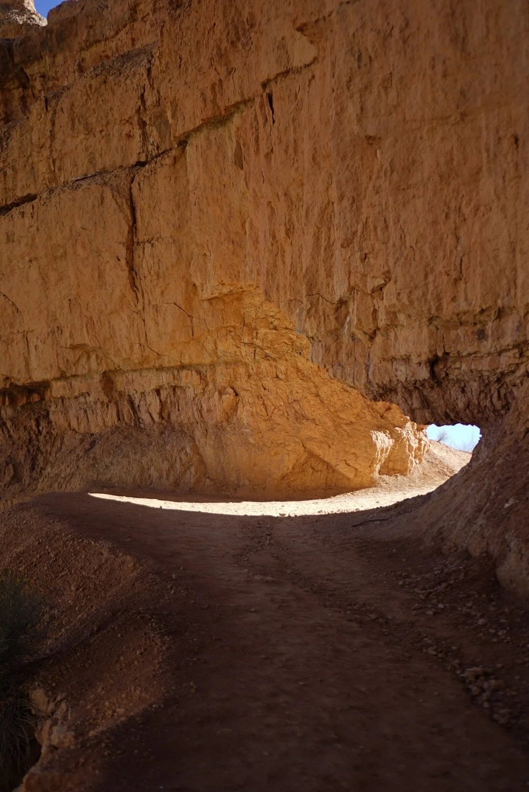 A view through a natural rock tunnel with orange and brown rock formations and a dirt path leading out to a bright sky.