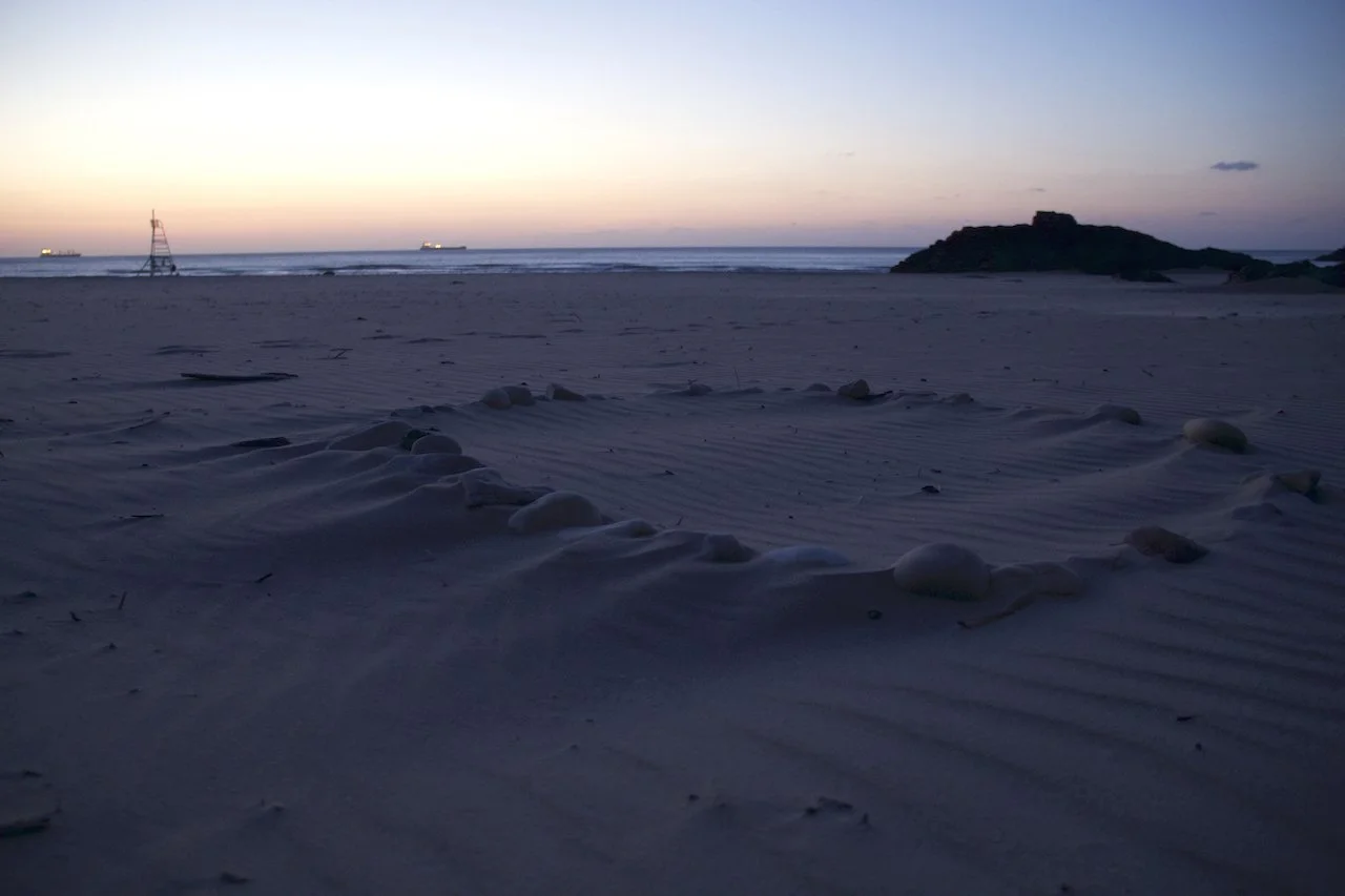 Beach at dusk with sand dunes, scattered rocks, and the ocean in the background, and ships visible on the horizon.