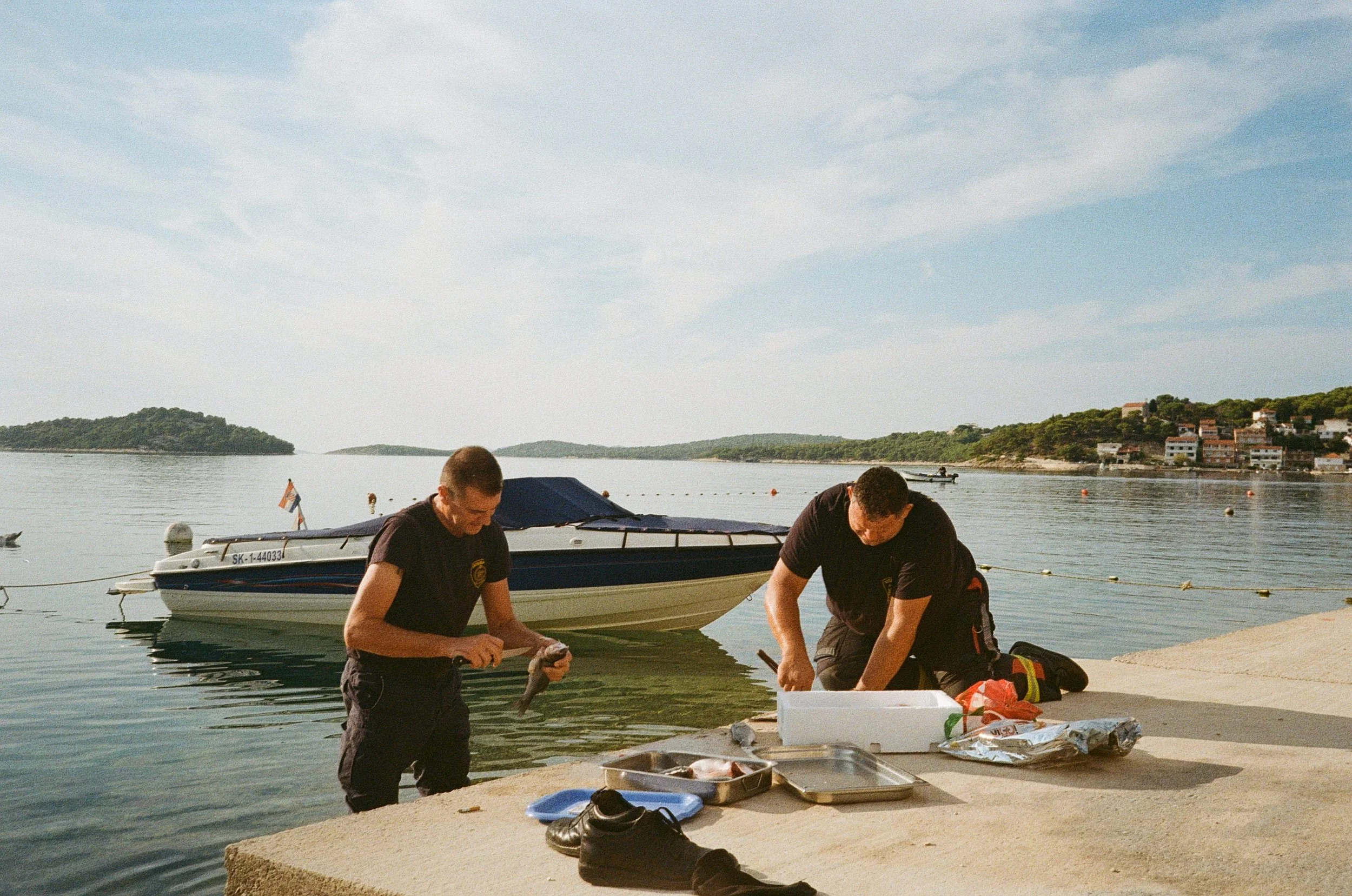 Two men cleaning fish at Tisno, Croatia, during a sunny sunrise. One man is holding a fish, and scattered fishing gear is visible on the dock. There are boats, a hillside with houses, and a calm body of water in the background.