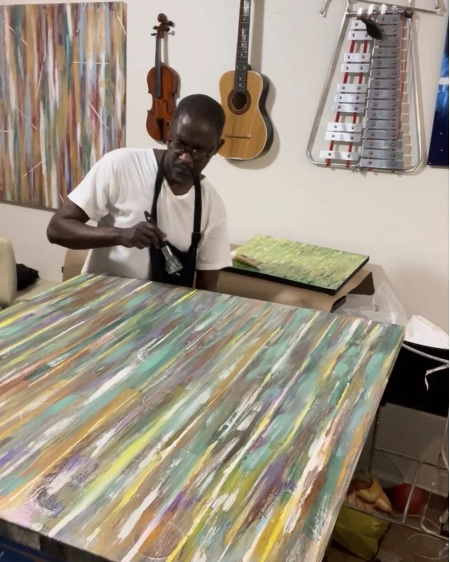 An man painting an abstract multicolored canvas in a room with musical instruments hanging on the wall and a xylophone.