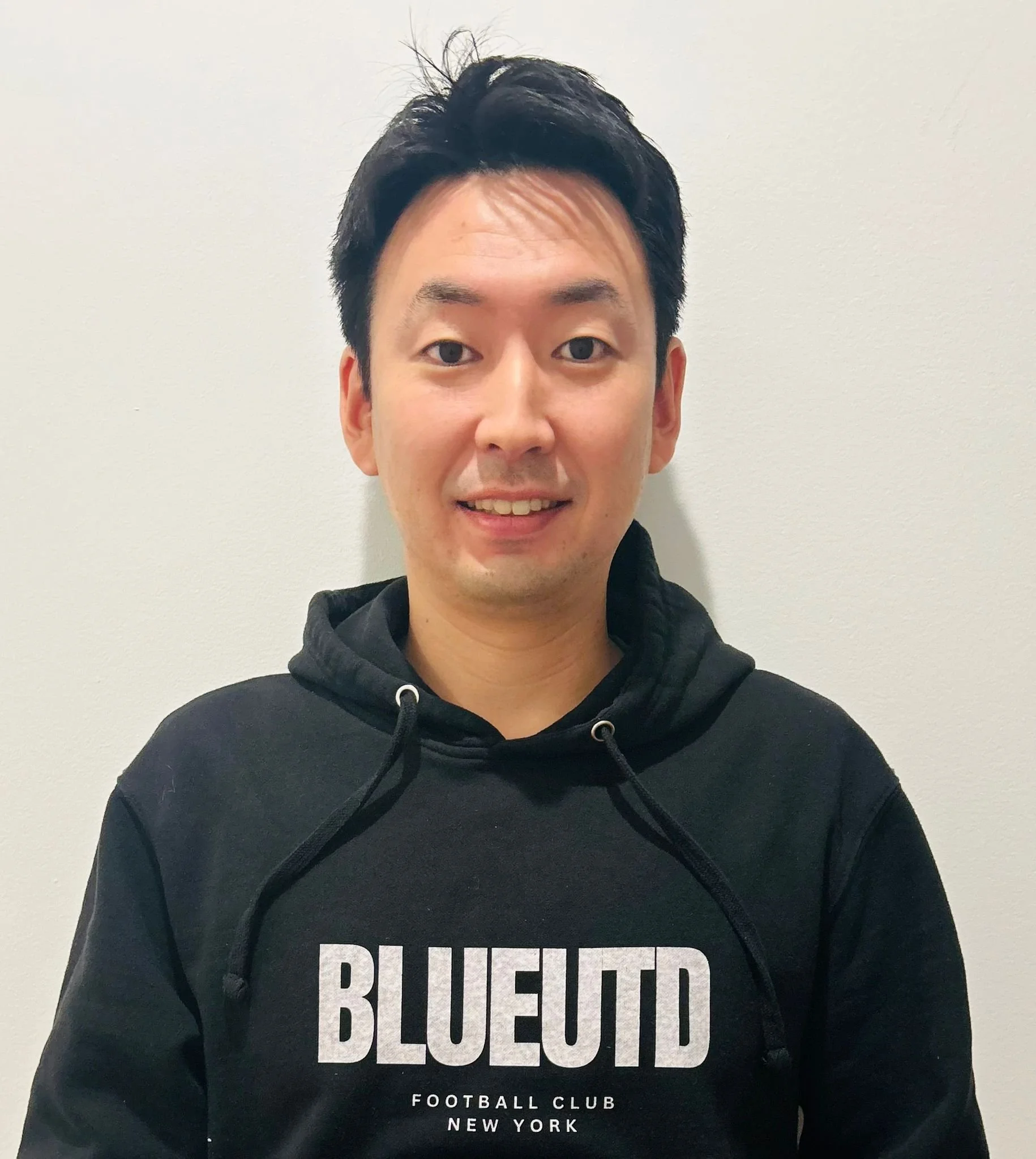 A man with black hair smiling, wearing a black hoodie with white text that reads 'BLUE TID' and 'FOOTBALL CLUB NEW YORK' against a plain, light-colored wall.