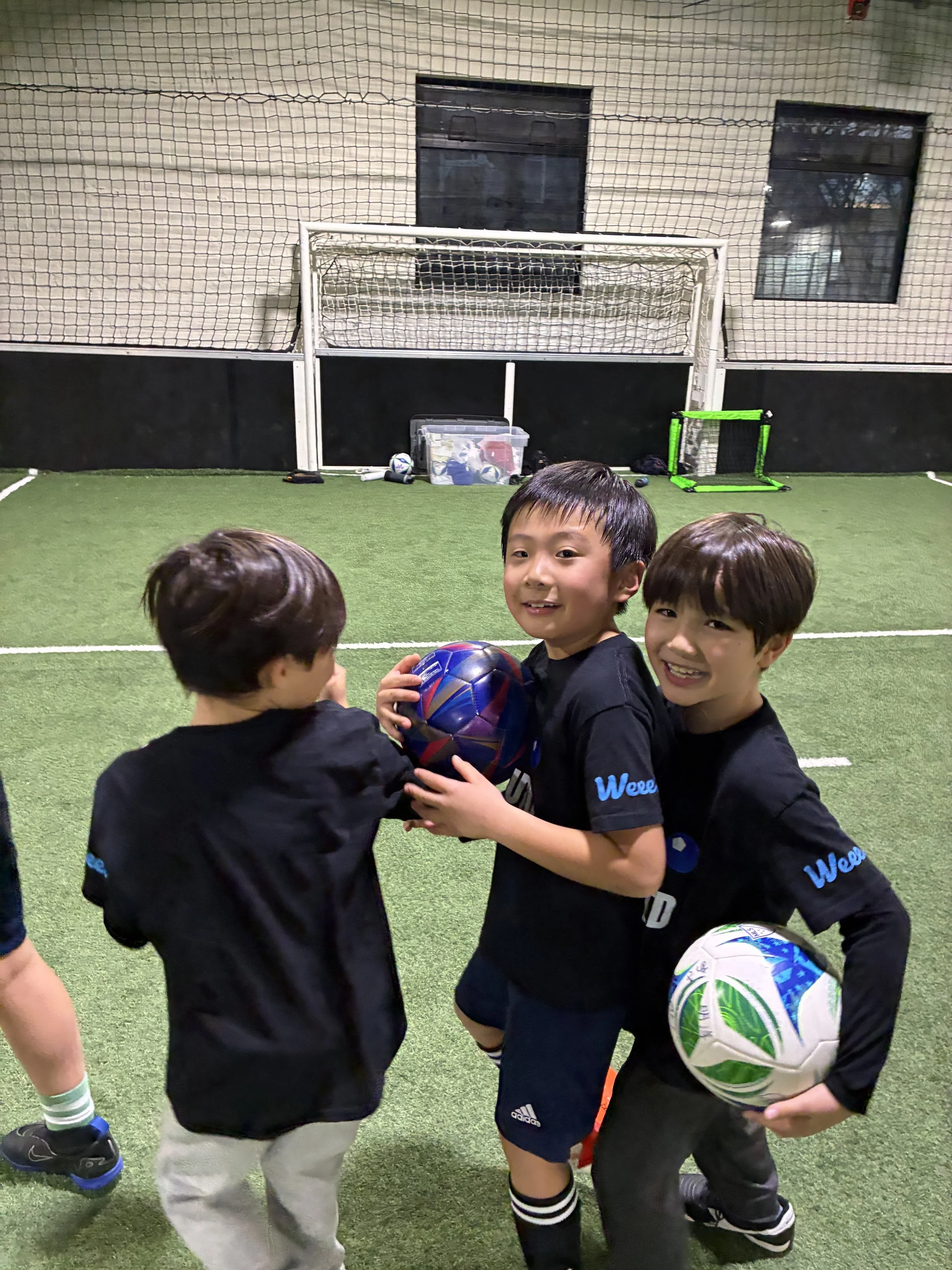 Three boys in sports jerseys holding soccer balls on an indoor turf field with a goal, netting, and sports equipment in the background.
