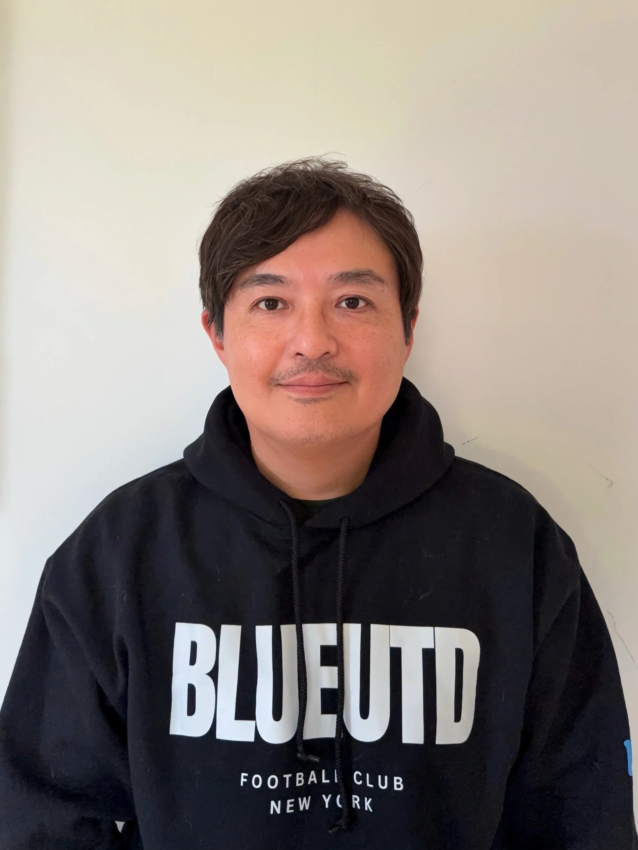 A man with short dark hair and light skin wearing a black hoodie with 'Blue Utd Football Club New York' written on it, standing against a plain white wall.