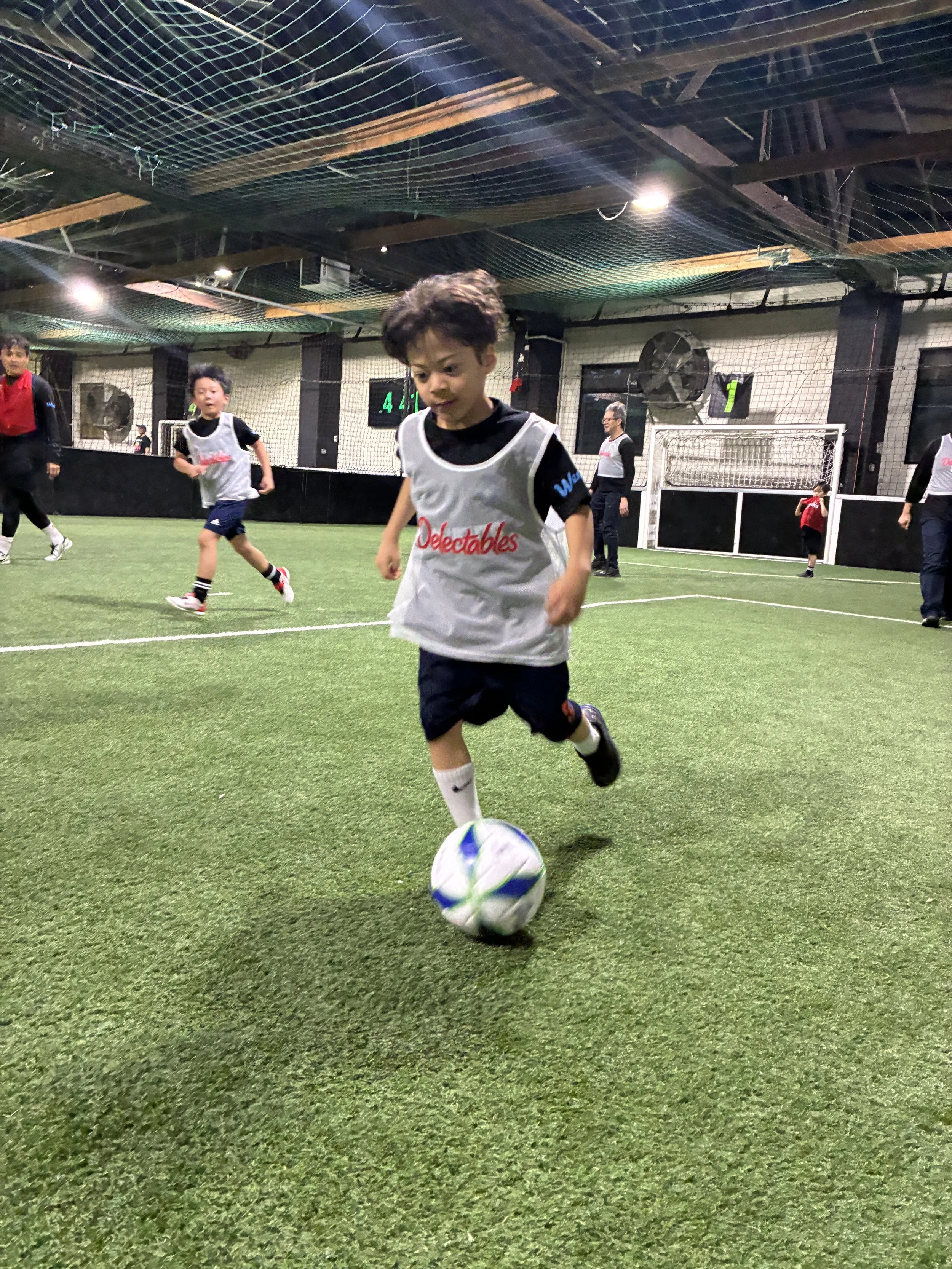 Young boy kicking a soccer ball on an indoor turf field with other children and adults in the background.