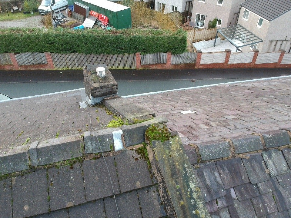 View from a rooftop showing a chimney with a cap, moss and dirt on roof shingles, and a street below with houses and a fence.