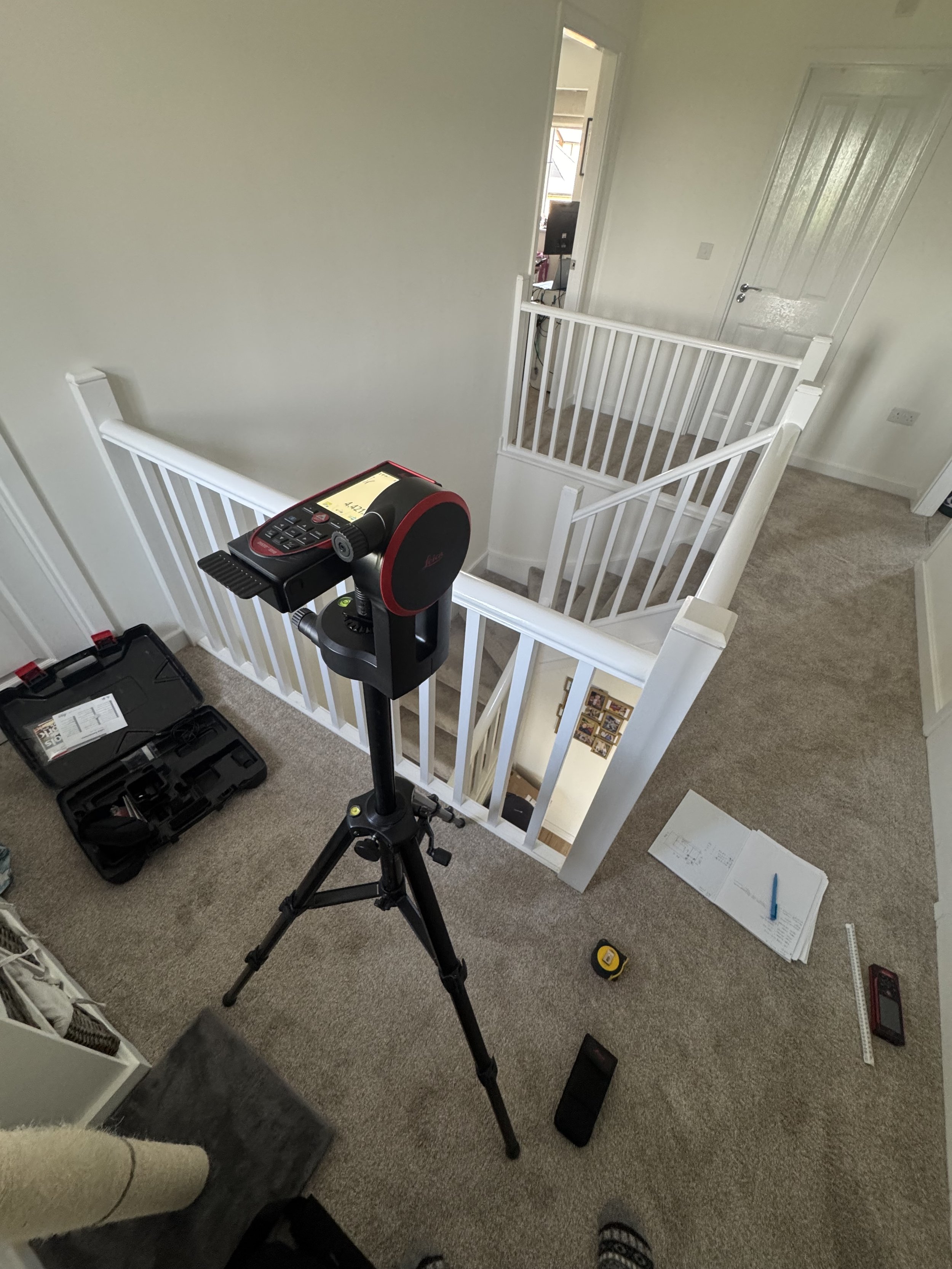 An indoor staircase landing with beige carpet, white railings, and a white door leading to another room. A treadmill with a red frame and black control panel is positioned nearby. There are open tool and equipment cases on the floor, papers, a tape measure, a spirit level, and a smartphone on the carpet.