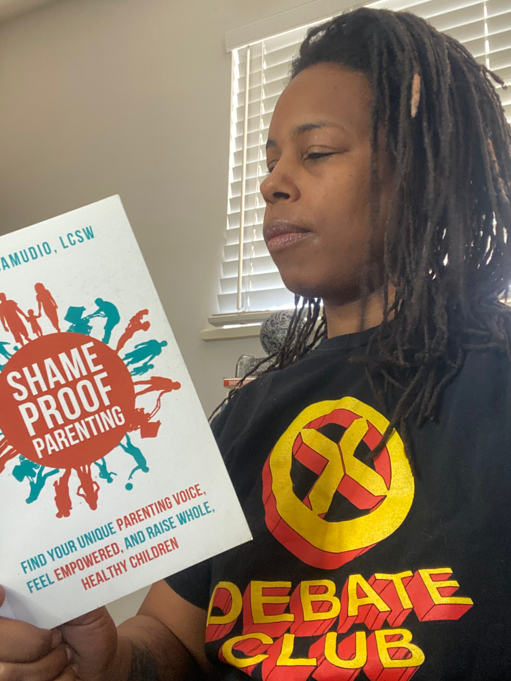 Dr. Mercedes Samudio reads her book Shame Proof Parenting, highlighting her work as an author and mental wellness advocate, while wearing a “X Debate Club” shirt in a softly lit indoor setting.