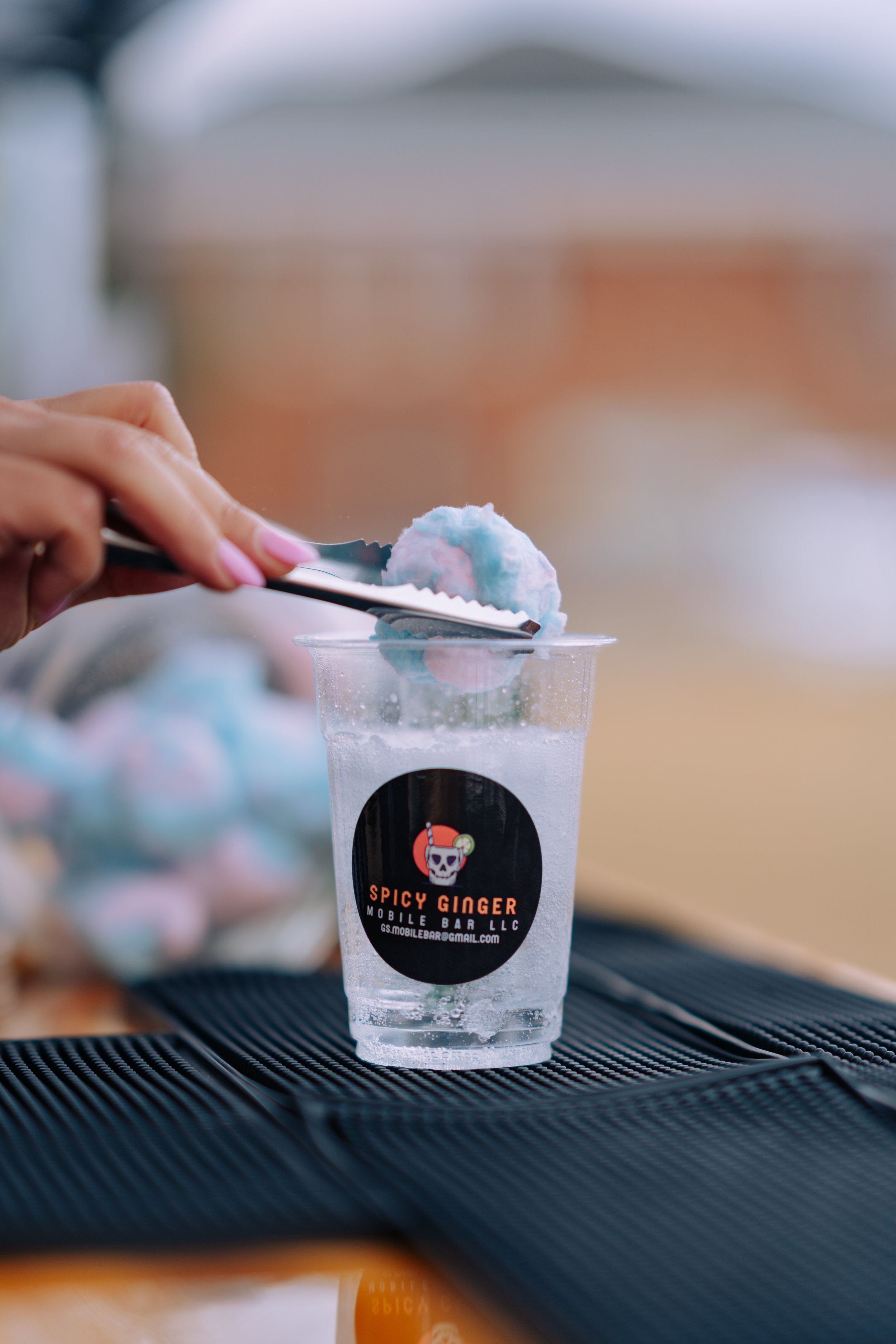 A hand with pink nails using tongs to pick up a blue and white cotton candy from a clear plastic cup on a black textured surface. The cup has a black circular logo with a skull and a red hat, with the words 'Spicy GINGER' and 'Mobile Bar LLC' written on it.