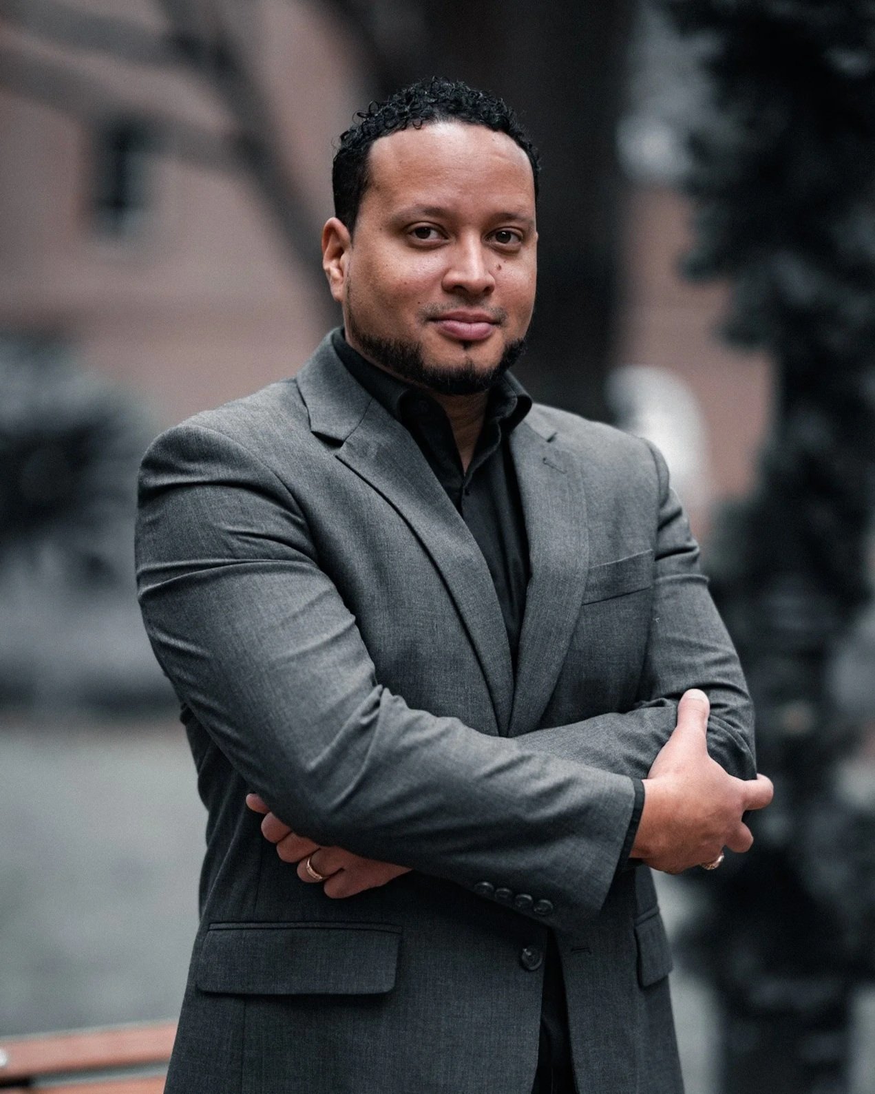 A man wearing a gray suit with a black shirt, standing outdoors with arms crossed, looking at the camera.