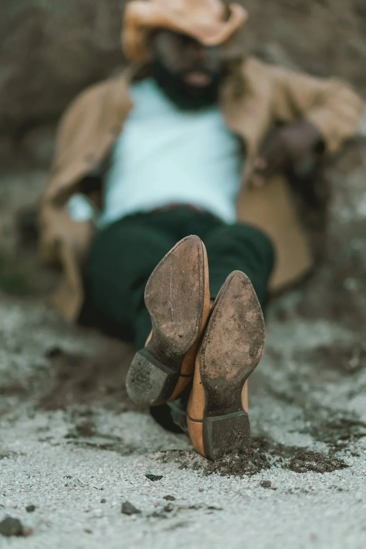 Person lying on ground with boots up, wearing a white shirt and tan jacket, on sandy terrain.