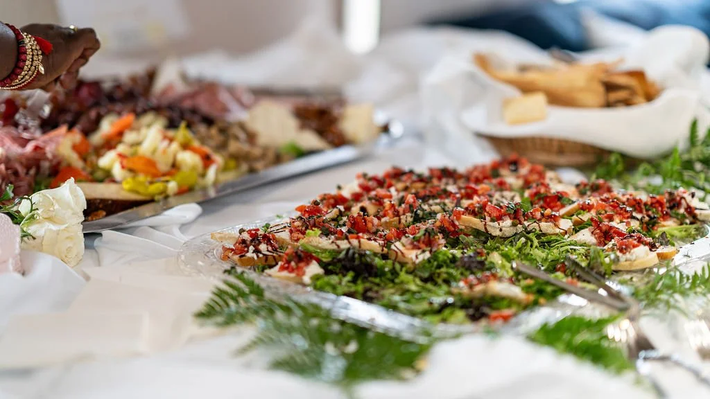 A buffet table with assorted dishes, including a tray of flatbread topped with greens, tomatoes, cheese, and herbs, and a platter of sliced meats and salad, decorated with greenery and flowers.
