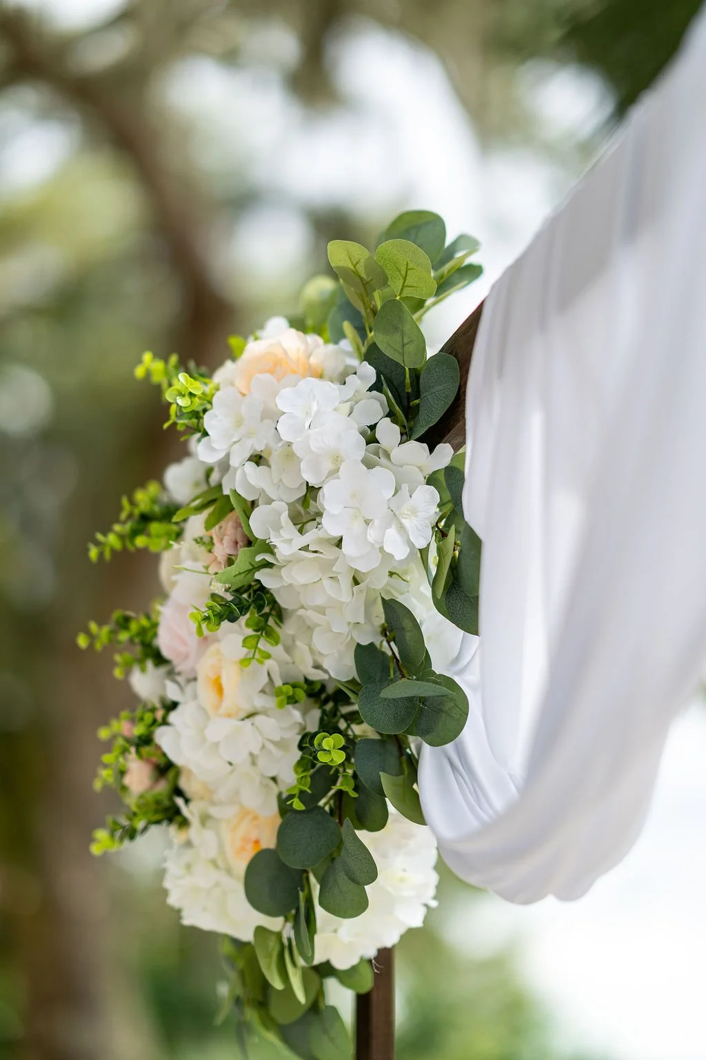 Close-up of a wedding floral arrangement with white hydrangeas and greenery on a white cloth backdrop.