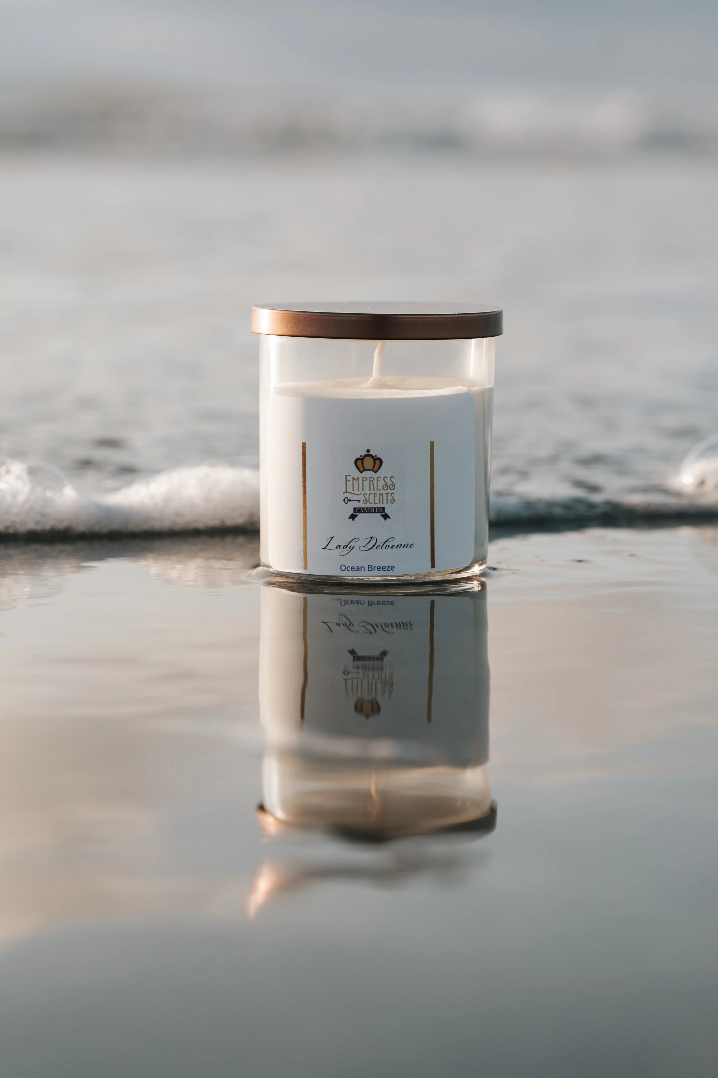 A white candle with a gold lid and a label that reads "Empress Scents, Lady Devonne, Ocean Breeze" reflecting on a calm water surface at the beach.