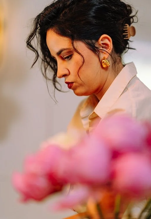 A woman with dark, curly hair styled in a loose updo, wearing gold earrings and a light-colored blouse, looking down thoughtfully with pink flowers in the foreground.