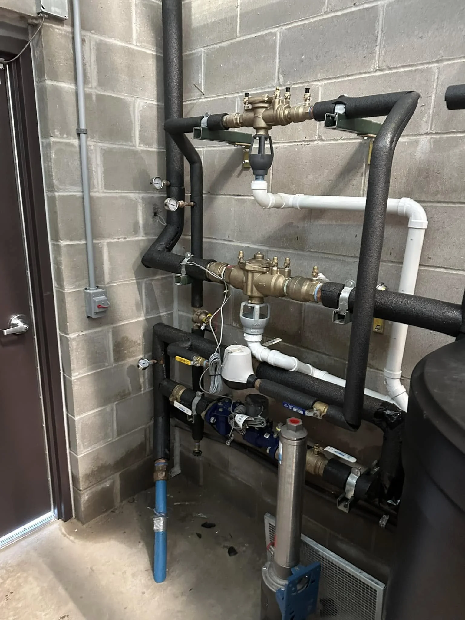 Industrial plumbing system with black and white pipes, gauges, and valves mounted on a concrete wall.
