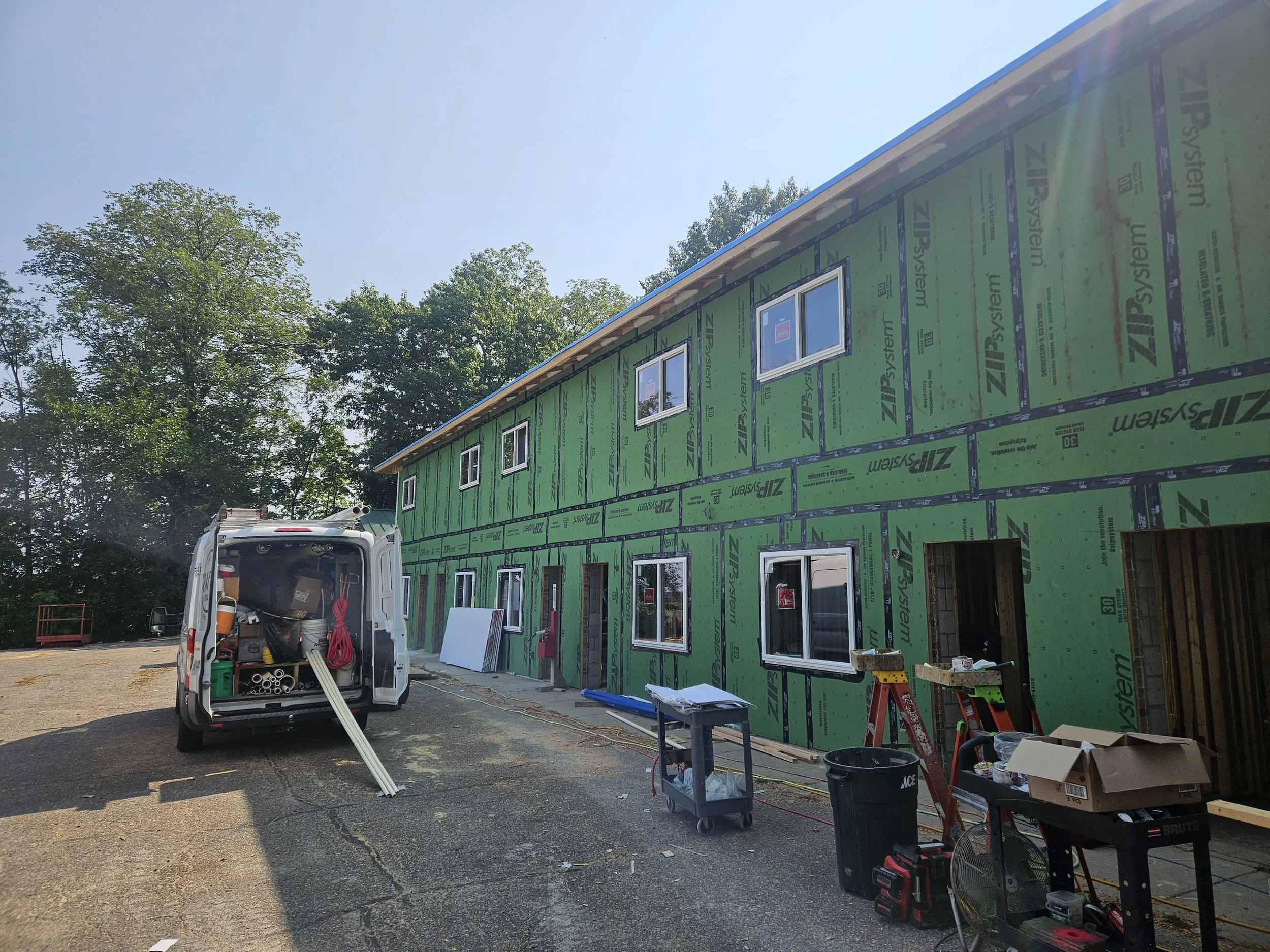 Exterior of a house under construction with green sheathing and several windows. Construction tools and supplies, including a white van with an open back, are visible in the foreground. Tall trees and a clear blue sky are in the background.
