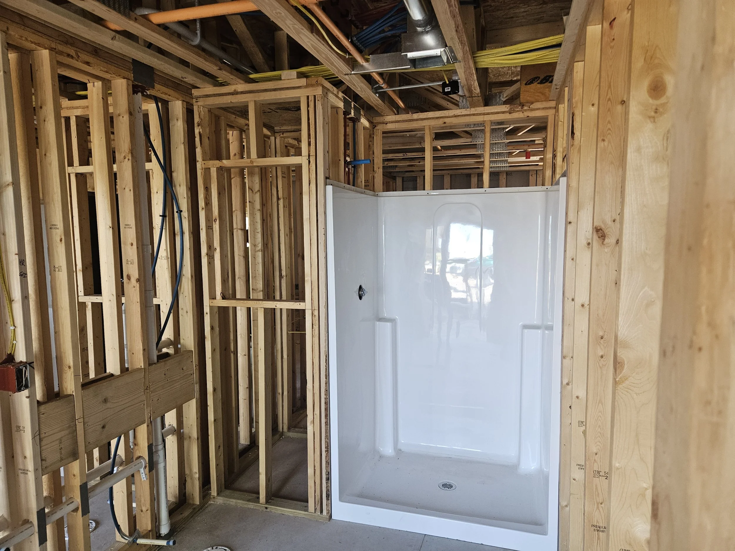 Construction site showing framed walls and a white shower base installed in a bathroom. Wooden studs and plumbing pipes are visible, indicating ongoing building work.