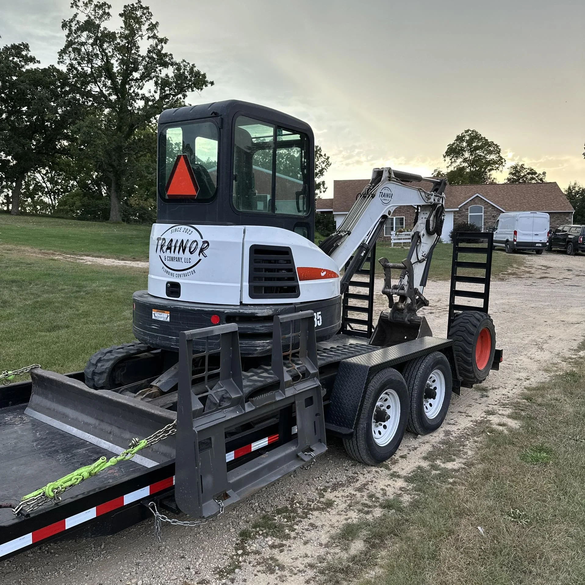 A construction mini excavator on a flatbed trailer parked on a grassy area near a residential house, with trees and a partly cloudy sky in the background.