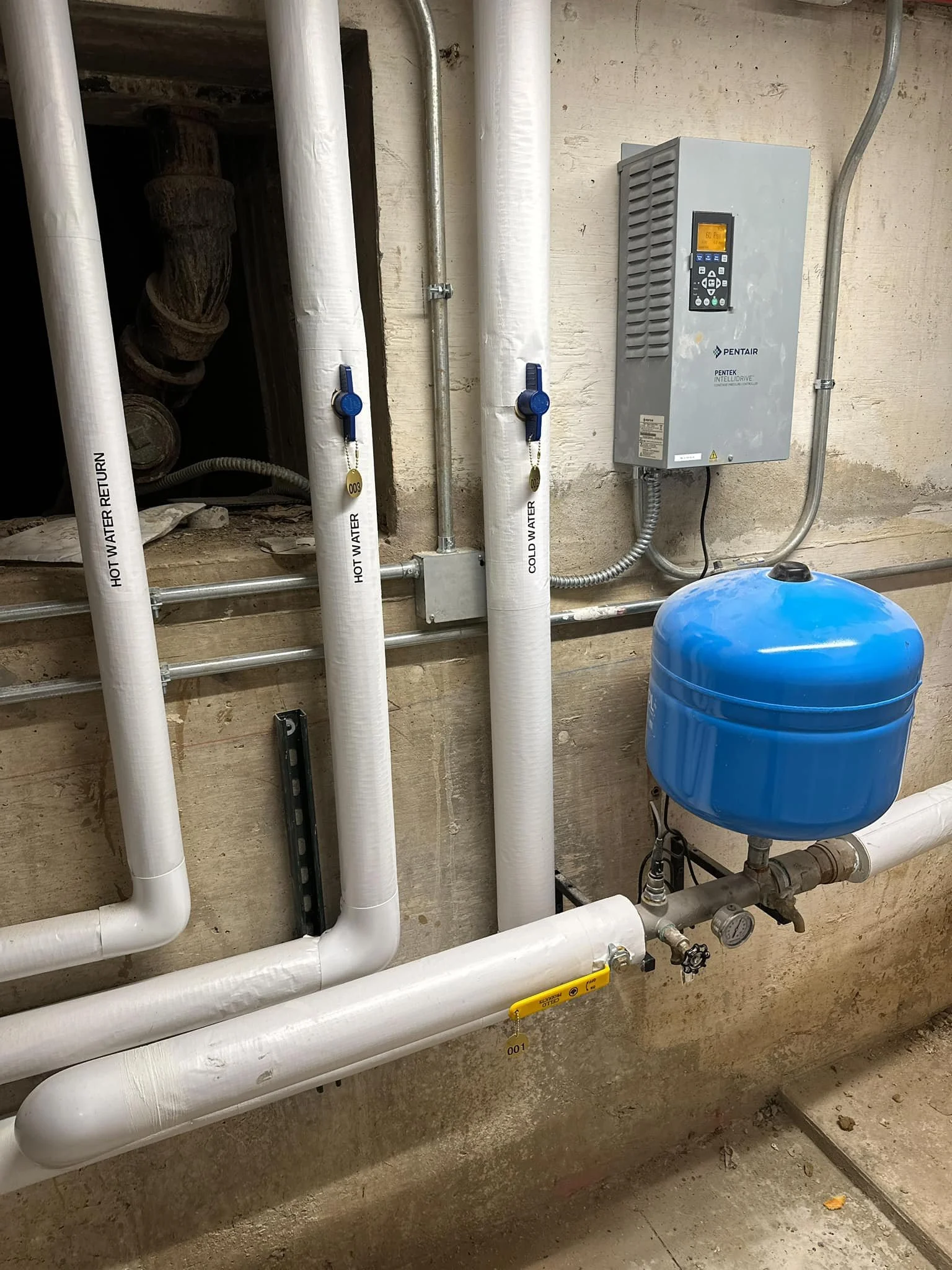 A plumbing system with multiple white pipes labeled for hot and cold water, a grey electrical box, and a large blue expansion tank, all mounted on a concrete wall.