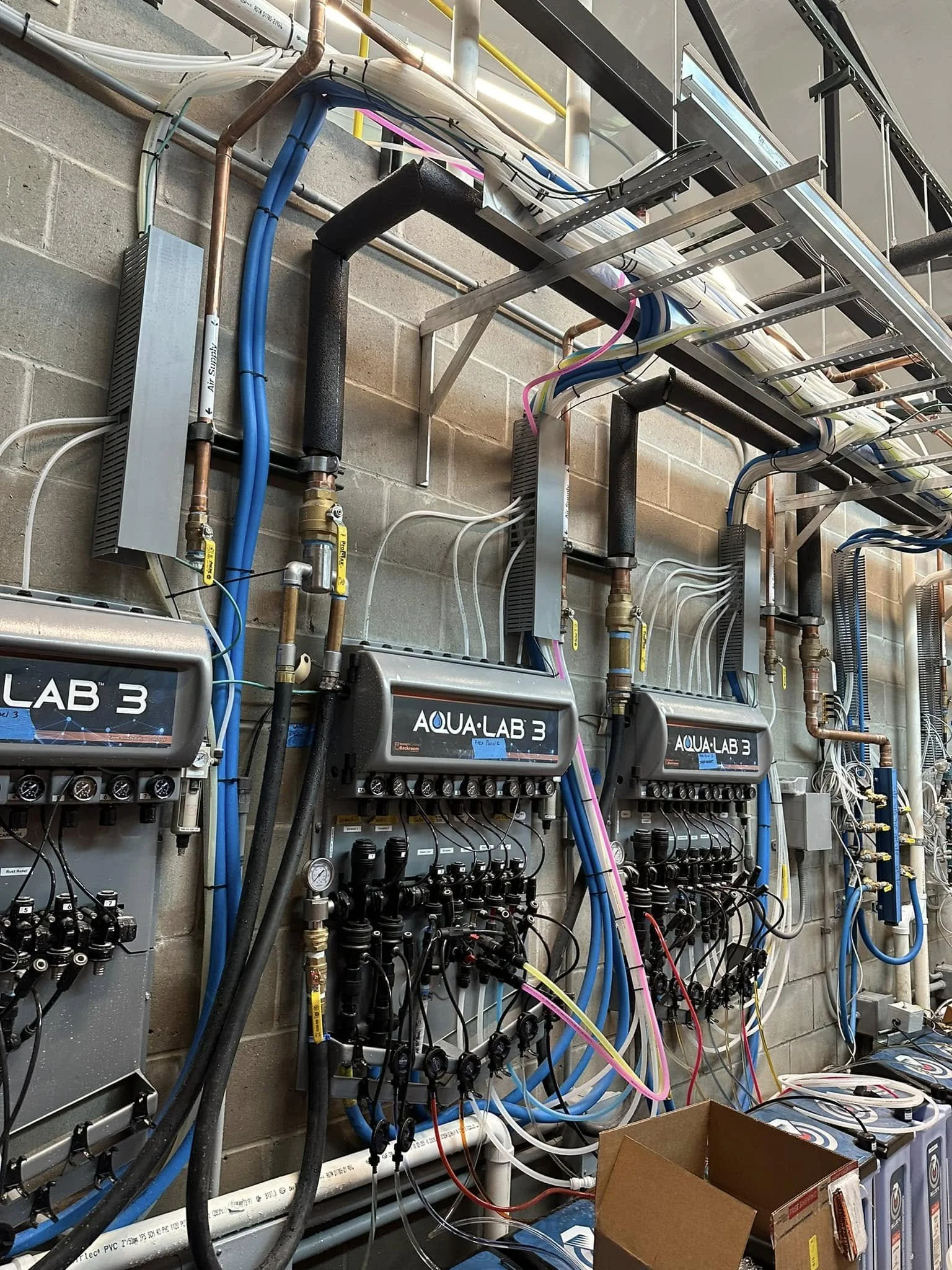 Multiple water control and distribution systems with pipes, valves, and pumps mounted on a concrete wall, labeled Aqua-Lab 3, in an industrial setting.