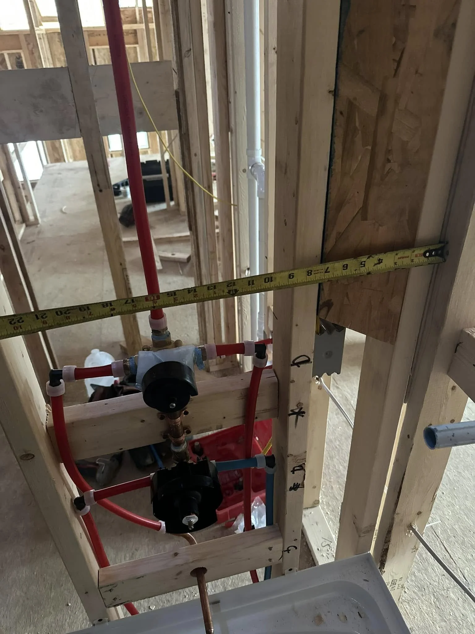 Inside view of a building under construction with exposed wooden framing, plumbing pipes, and measuring tape.