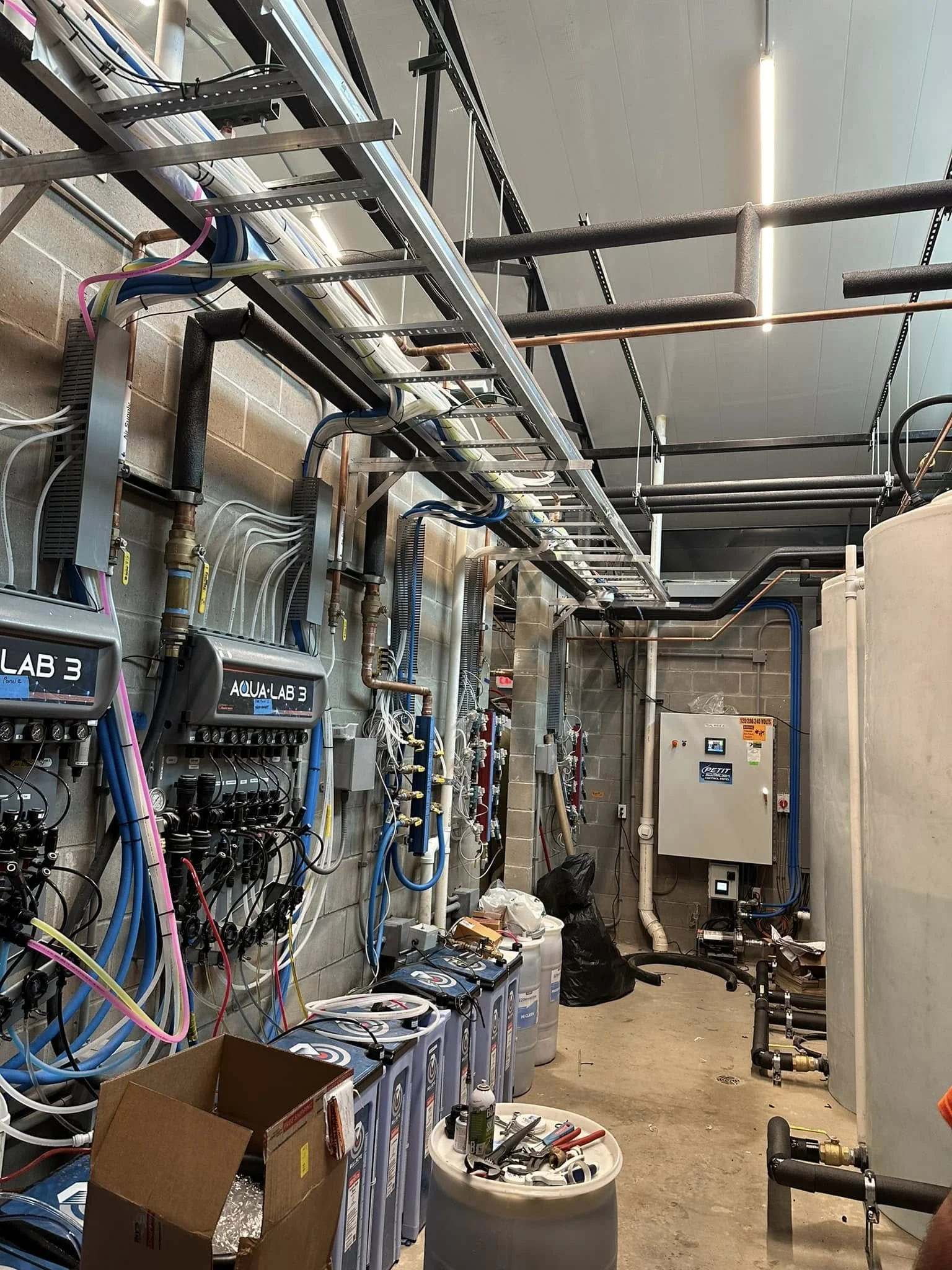 A utility or mechanical room with pipes, electrical wiring, and equipment for water or HVAC systems. There are labeled devices, pipes, tubing, and tools, indicating ongoing installation or maintenance work.