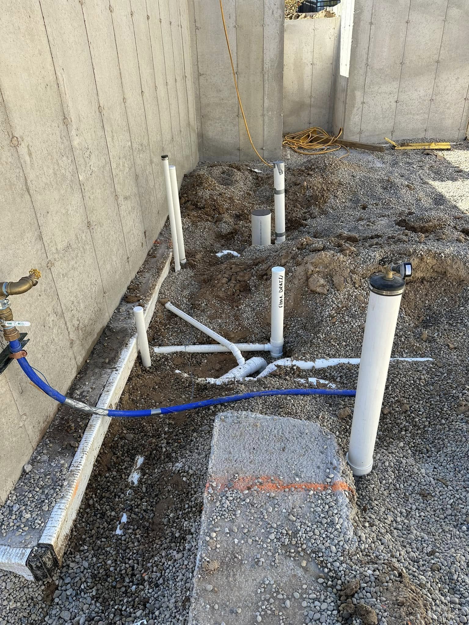 Construction site with exposed pipes and gravel ground near a concrete wall, with yellow extension cords on the ground.