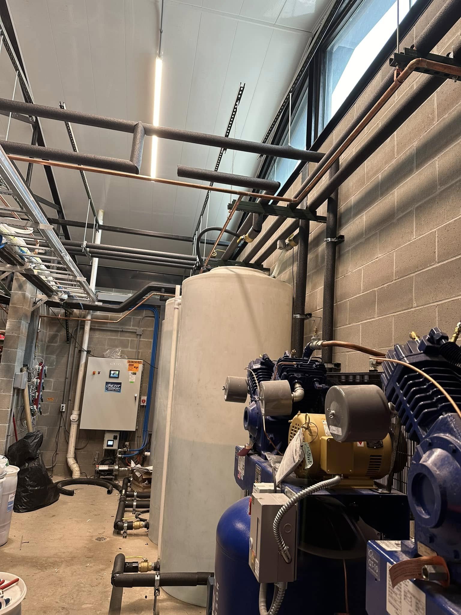 An industrial room with machinery, pipes, and equipment for water or air filtration, including a large white tank, a blue tank, and various black and copper pipes, with a window on the wall and a control panel on the left side.