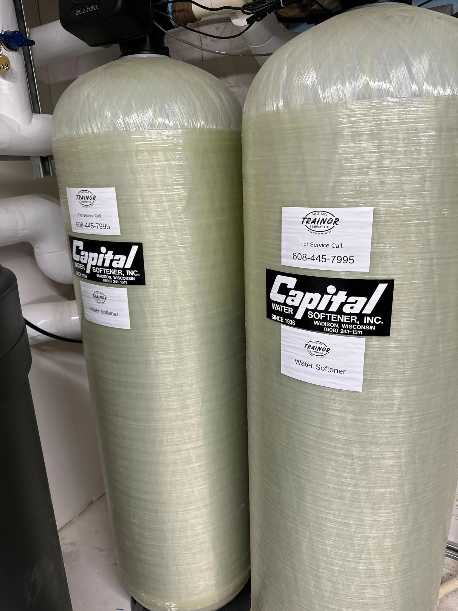 Two large water softener tanks with labels from Capital Water Softener, Inc. and Trainor & Company, LLC, placed in a utility room, connected to white pipes and other equipment.